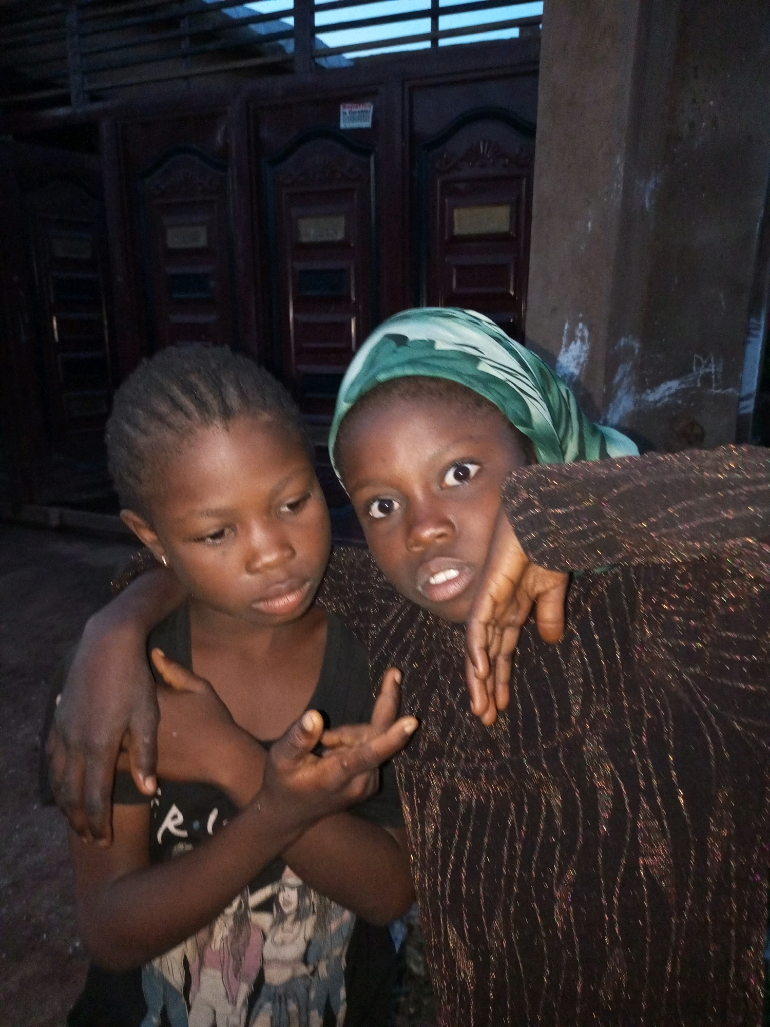 The image depicts two young girls in what appears to be an outdoor setting at dusk, with a building featuring dark wooden doors in the background. The girl on the left, wearing a black t-shirt, has her left arm around the other girl and her right arm across her chest. She has small braids in her hair and a small gold earring. The girl on the right is wearing a green headscarf and a dark, sparkly patterned garment. Her right hand is resting on her cheek, and her left hand is near the other girl's arm. Their expressions suggest a moment of connection or shared experience. The environment appears somewhat urban, possibly a residential area in the city of Tempure, Nigeria, given the building's architecture and the lighting conditions.