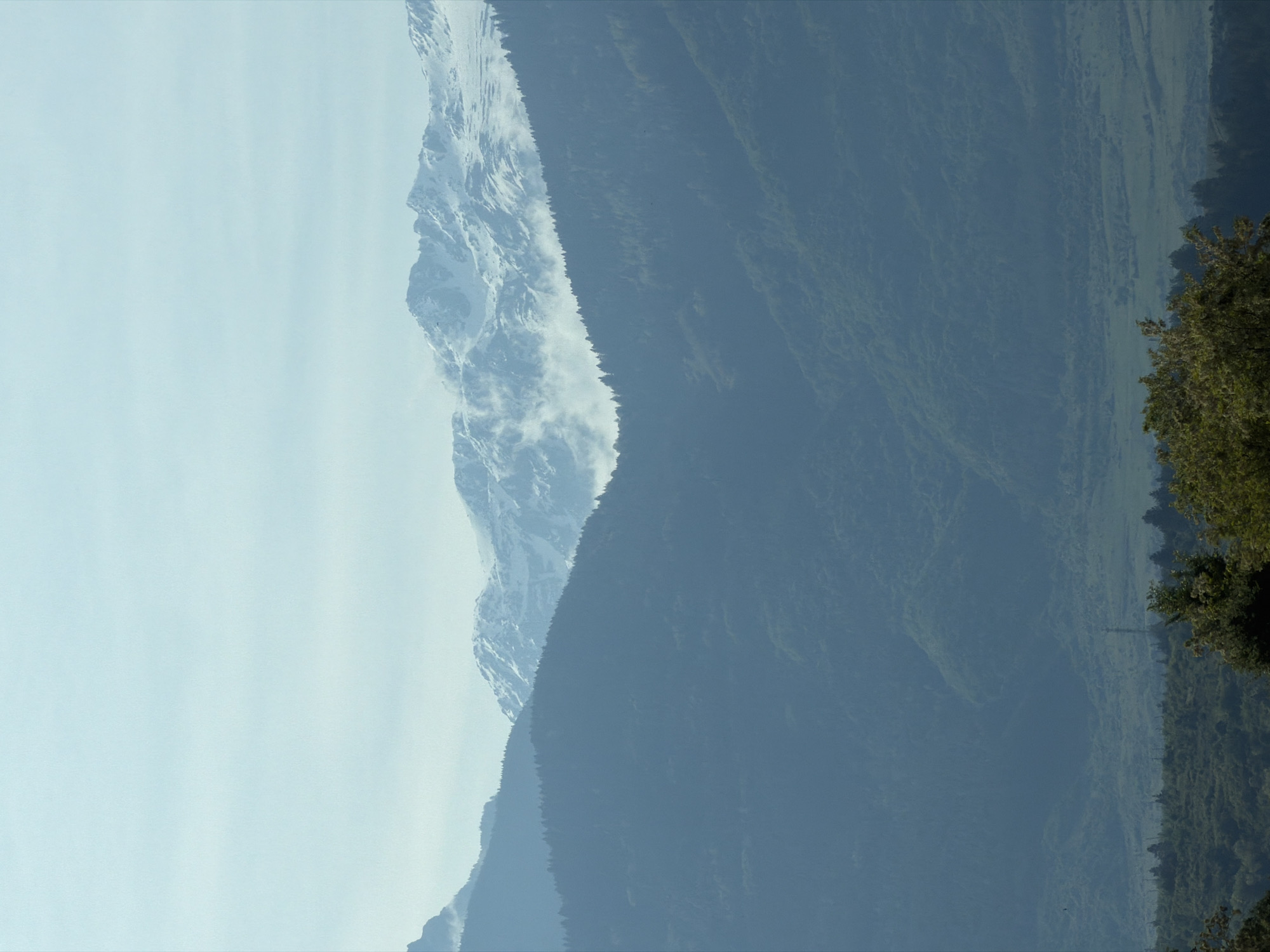 The image shows a mountain range, likely in Bulgaria, based on the implied location in Balanovo. The left side of the image features a snow-capped mountain with visible rock formations. The right side reveals a densely forested mountainside, possibly indicating lower elevations. The sky appears light, suggesting a clear or slightly hazy day.