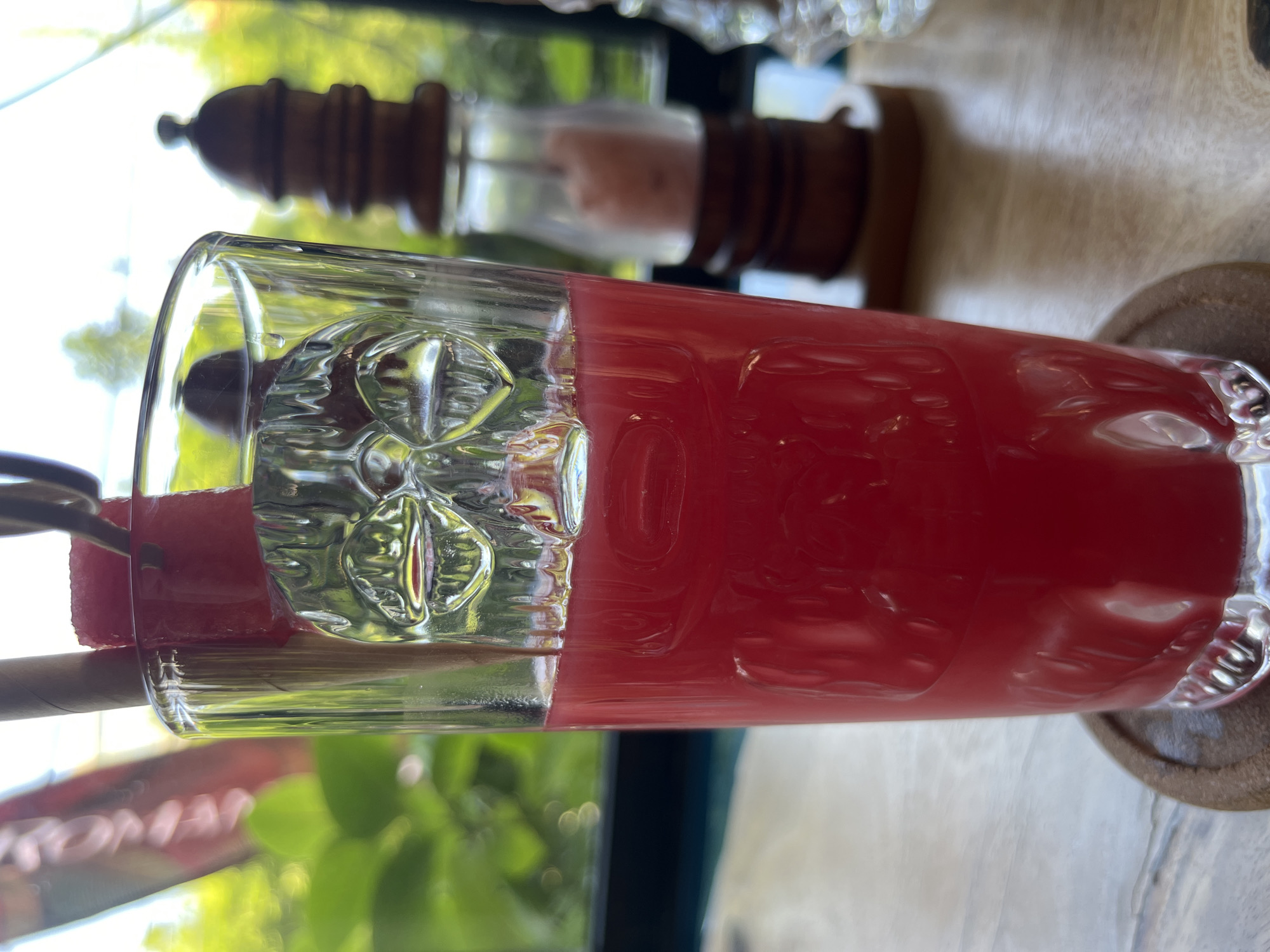 A tall glass of red liquid, likely watermelon juice, sits on a round, brown coaster on a light-colored wooden table. The glass has a patterned texture in its upper half. A slice of watermelon is attached to the glass's rim. A paper straw is also present. In the background, there is a blurred outdoor scene with greenery. Behind the glass, there is a salt and pepper shaker set. This image was likely taken in North Kuta, Indonesia.