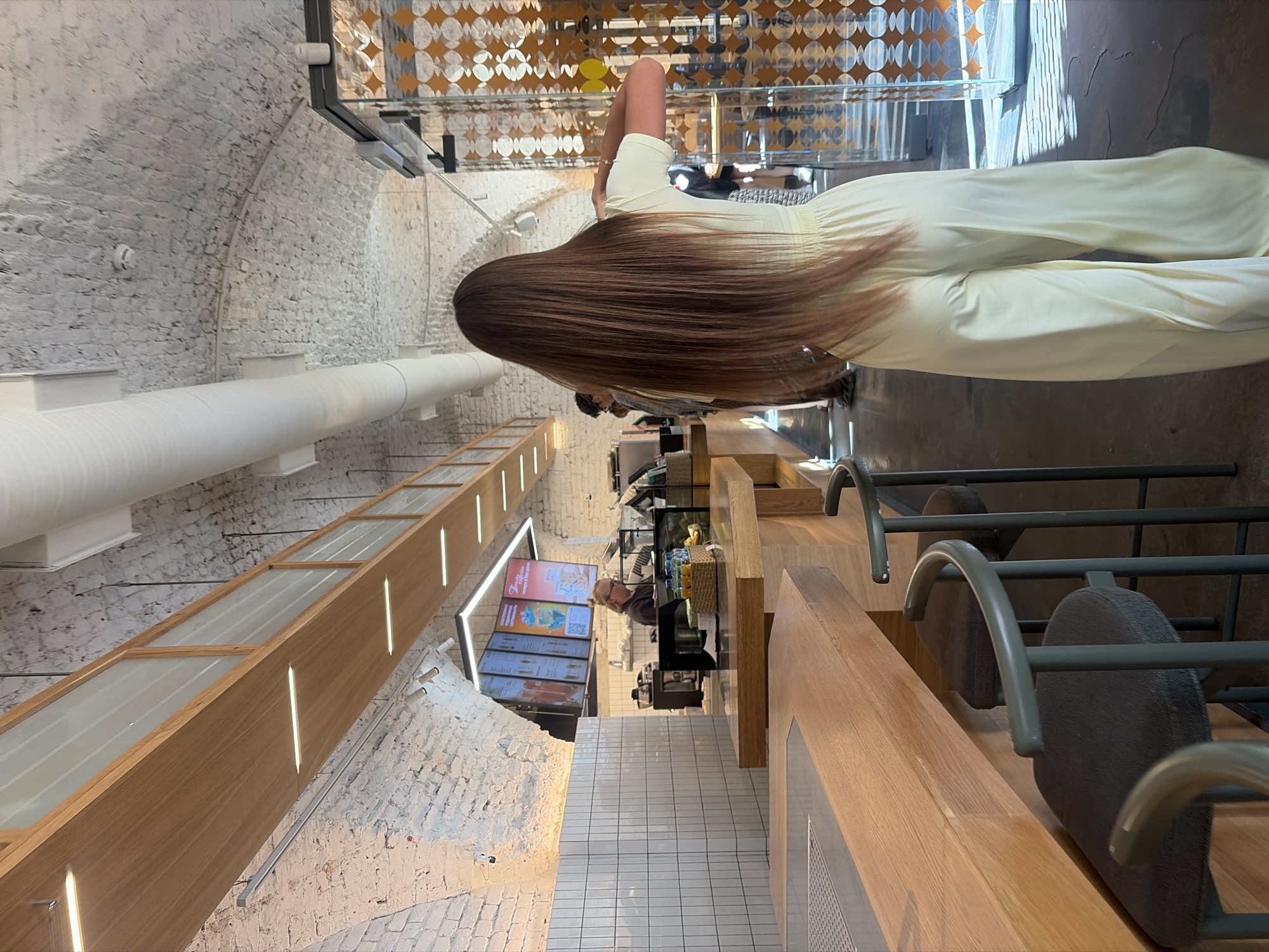 The image depicts the interior of a modern cafe located in Saint Petersburg, Russia. The cafe features a long wooden counter with several dark-colored stools. The walls and ceiling are constructed of white brick, with visible ventilation pipes and lighting fixtures. In the background, a barista stands behind the counter, serving customers. A woman with long brown hair, wearing a light yellow jumpsuit, is standing in the foreground, facing away from the camera. The cafe appears to be clean and well-lit, with a relaxed atmosphere.