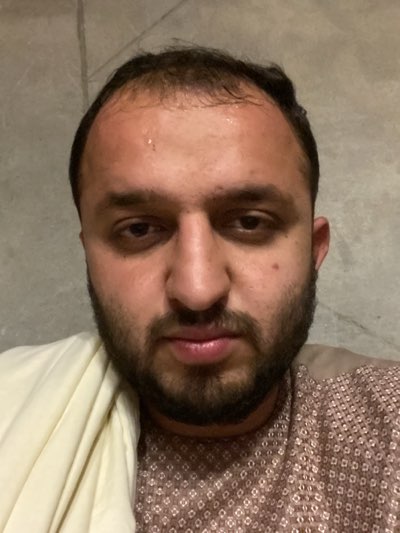 A close-up, straight-on portrait shows a man with a dark beard and short, dark hair. He has brown eyes and a light skin tone. The man appears to be sweating slightly on his forehead. He is wearing a patterned brown and tan shirt and a light-colored shawl draped over one shoulder. The background is a plain, light gray wall. There are no indicators of the location being Kandahar, Afghanistan.