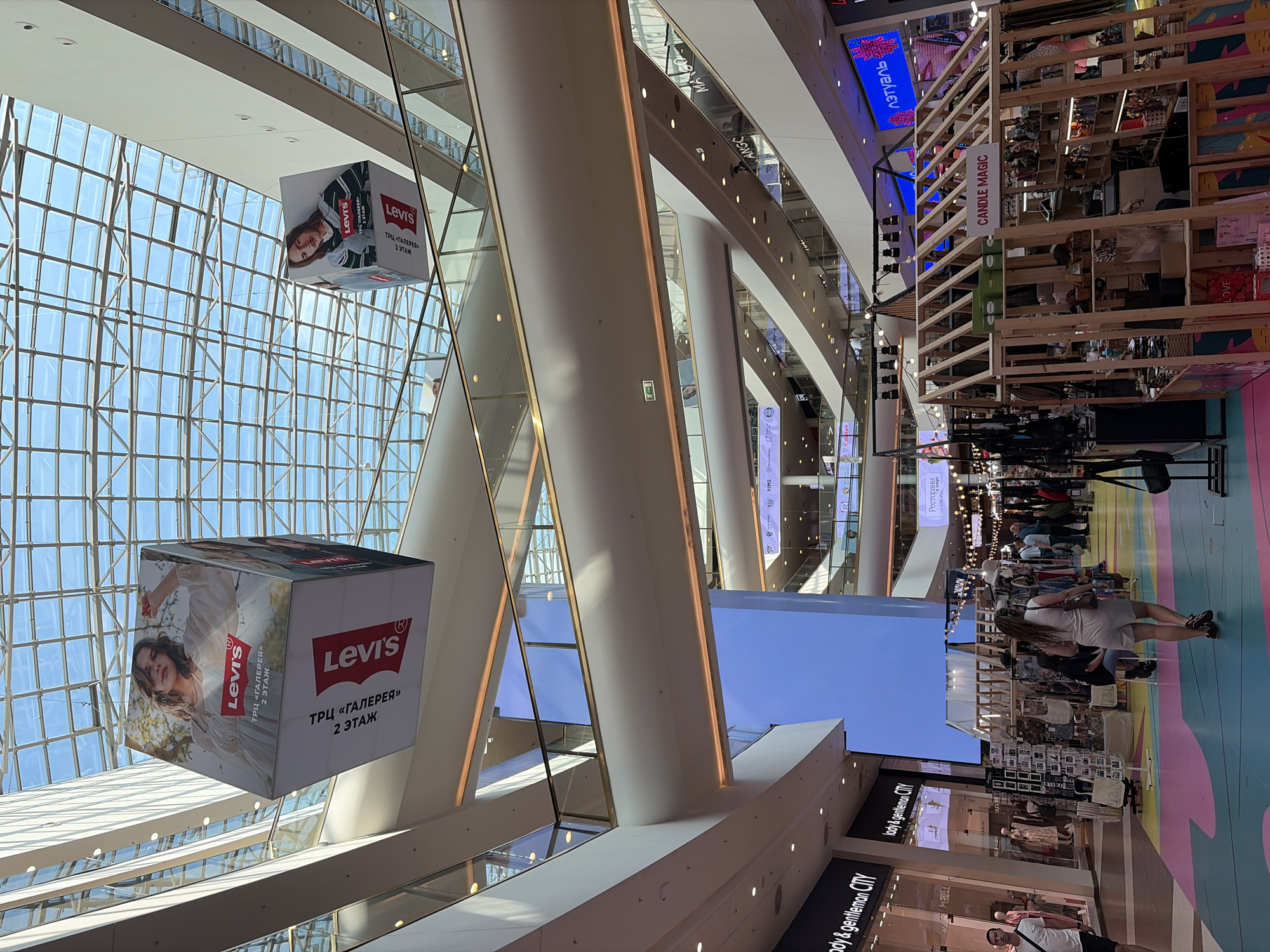 This media file captures the interior of a brightly lit, modern shopping mall, likely in Saint Petersburg, Russia. The architecture features a large glass ceiling that allows natural light to stream in, illuminating the multi-level structure. Large, white cylindrical columns support the upper floors.
Dominating the view are two large, cube-shaped advertisements suspended from the ceiling. Both feature the iconic Levi's logo and imagery of a young woman. The text on the advertisements indicates "ТРЦ «ГАЛЕРЕЯ»" and "2 ЭТАЖ," suggesting they are promoting the Levi's store on the second floor of the Galeria shopping center.
Further into the mall, various shops and displays are visible. One prominent display, constructed from light-colored wood, is labeled "CANDLE MAGIC." Another sign reads "лэтуаль," a well-known Russian cosmetics and perfume retailer. People can be seen browsing in the background, suggesting a normal day of shopping. The overall impression is one of a busy and vibrant retail environment. The natural light and clean lines of the mall architecture contribute to a sense of spaciousness and modernity.