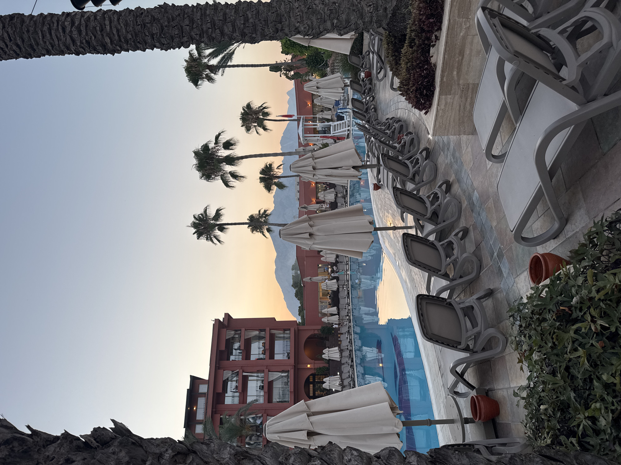 This image depicts an outdoor swimming pool area at a resort in Kemer, Turkey. The scene is set during sunset, with the sky displaying hues of orange and yellow, suggesting a warm and pleasant evening. Palm trees are scattered throughout the area, adding to the tropical ambiance.
The main subjects are the swimming pool, lounge chairs, umbrellas, and a multi-story building that appears to be part of the resort. The pool is a vibrant blue, reflecting the surrounding structures and the sky. Numerous lounge chairs, mostly made of grey plastic, are arranged around the pool, some occupied by people, indicating leisure activity. Several closed white umbrellas are positioned strategically for shade. The building, painted in a reddish-brown hue, features multiple balconies and windows, suggesting guest rooms.
In the background, mountains are visible, partially shrouded in the soft light of the setting sun. The overall mood conveyed is one of relaxation and tranquility, characteristic of a holiday destination. The time of day is clearly evident from the warm glow in the sky and the long shadows cast by the palm trees and loungers. The weather appears clear and calm.
There is no visible text in the image. The location context provided specifies Kemer, Turkey, which aligns with the visual cues of palm trees, a resort-style setting, and mountainous terrain often found in the region.