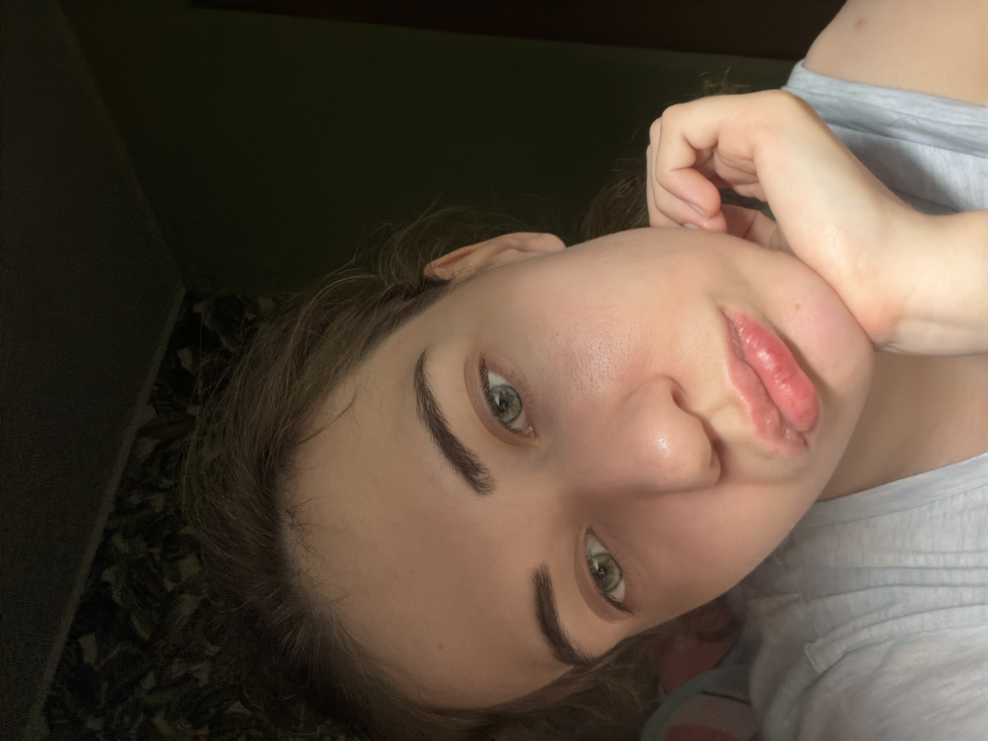 A close-up, low-angle, selfie-style photograph shows the face of a young woman with light brown hair and blue-green eyes. She is resting her chin on her right hand, which is positioned near her jawline. Her lips are slightly parted and appear to be rouged. The lighting suggests an indoor environment, possibly a bedroom, with a patterned wallpaper visible in the upper left corner and a dark wall in the background. The woman is wearing a light blue t-shirt. The image does not contain any specific landmarks or clues to pinpoint its location within Zavodoukovsk, Russia.