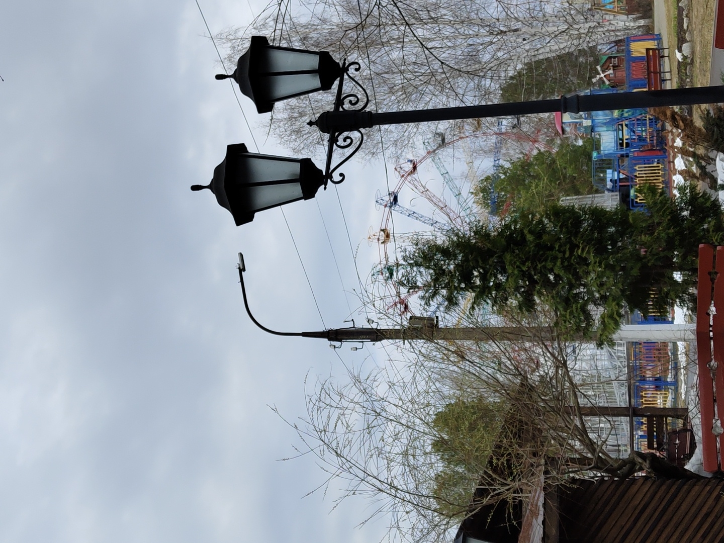 The image depicts a scene in an urban environment, likely a park or recreational area. In the foreground are two ornate black lampposts, classic in design, which stand out against the overcast sky. Further into the scene, a modern streetlight with a curved arm is visible.
In the background, there's a large Ferris wheel, indicating the presence of an amusement park or fairground. Colorful structures, possibly playground equipment or buildings associated with the park, can also be seen. Trees and foliage are interspersed throughout, adding a natural element to the urban landscape. The bottom of the image features the edge of a building, possibly a concession stand or park facility, constructed with corrugated metal.