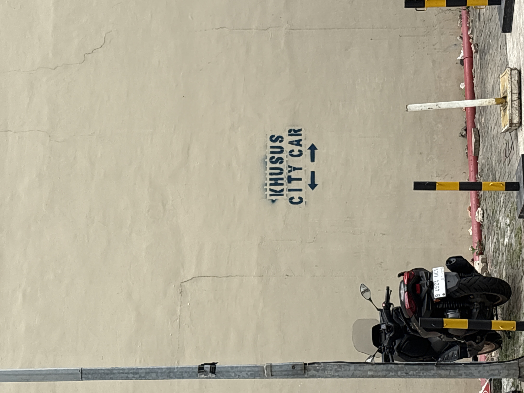 The image shows a wall in North Jakarta, Indonesia, with a stencil spray-painted onto it that reads "KHUSUS CITY CAR" and features two arrows pointing in opposite directions. To the left of the wall, a motorcycle is parked. The wall itself is a light beige color and appears to be made of stucco or concrete, showing some signs of wear and tear, including visible cracks. In the foreground, there are several bollards, some black and yellow striped, and one white. A red pipe runs along the base of the wall, with scattered debris and rocks on the ground.