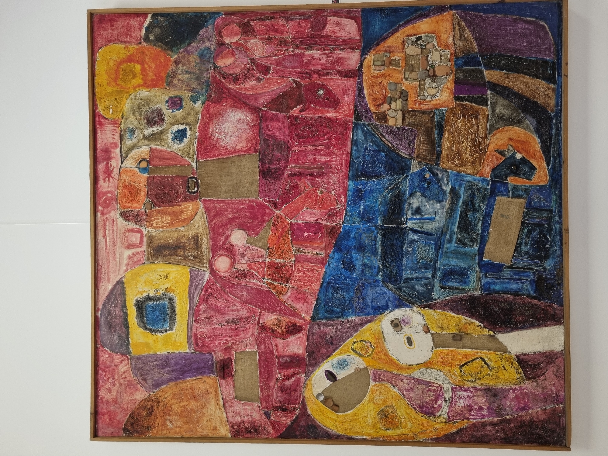 The image is a photograph of an abstract painting in a wooden frame. The painting features a collage of shapes and textures in various colors, including red, blue, yellow, brown, and purple. Some shapes are filled with stones. There are no people present. The painting has been photographed in a setting with white walls. The location of Elena, Bulgaria, is not explicitly discernible from the image.