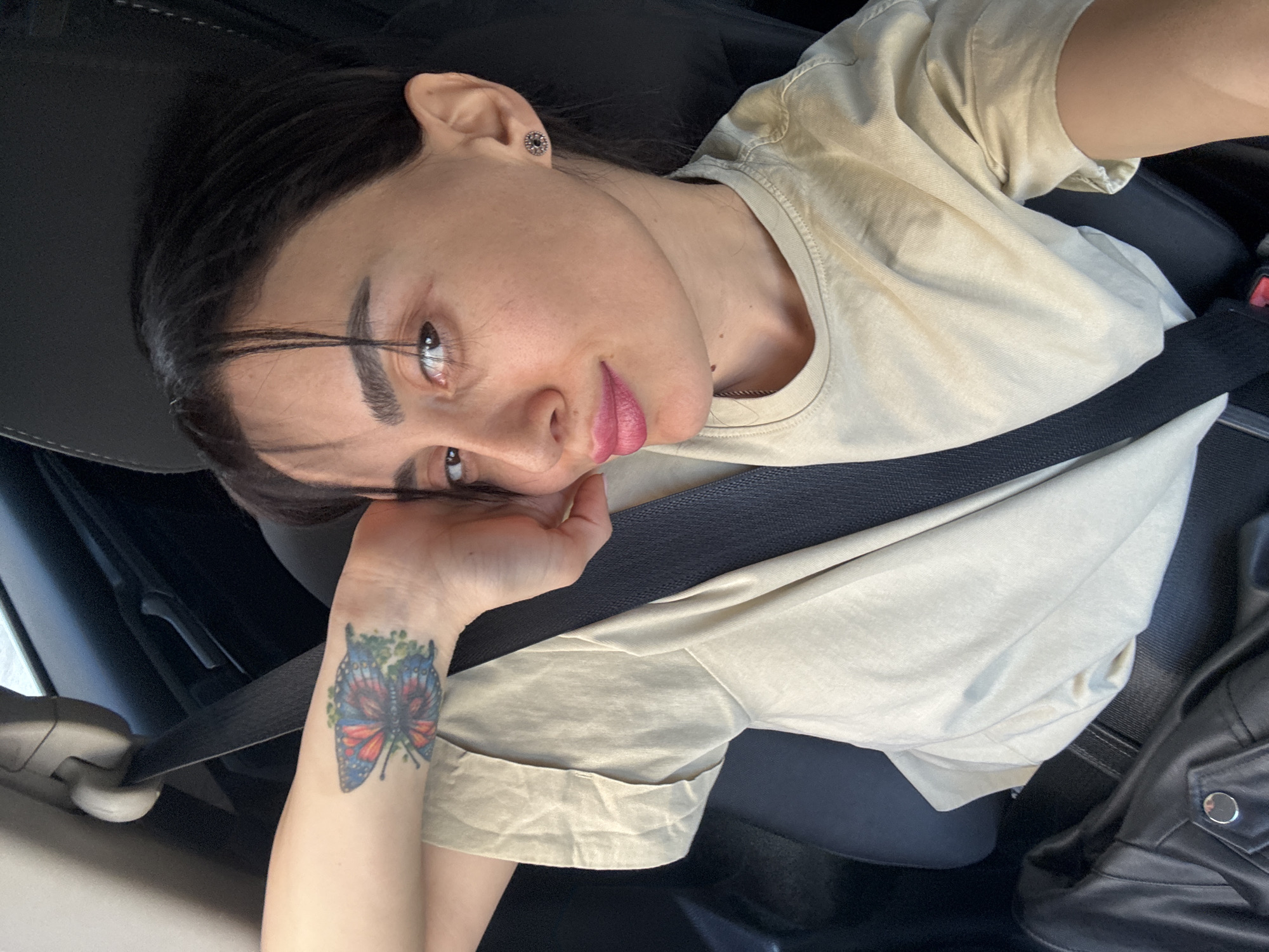 A woman with dark hair and light eyes is in a car, wearing a tan T-shirt and a seatbelt. She has a butterfly tattoo on her left wrist. The interior of the car is black. The location is not discernible from the image.