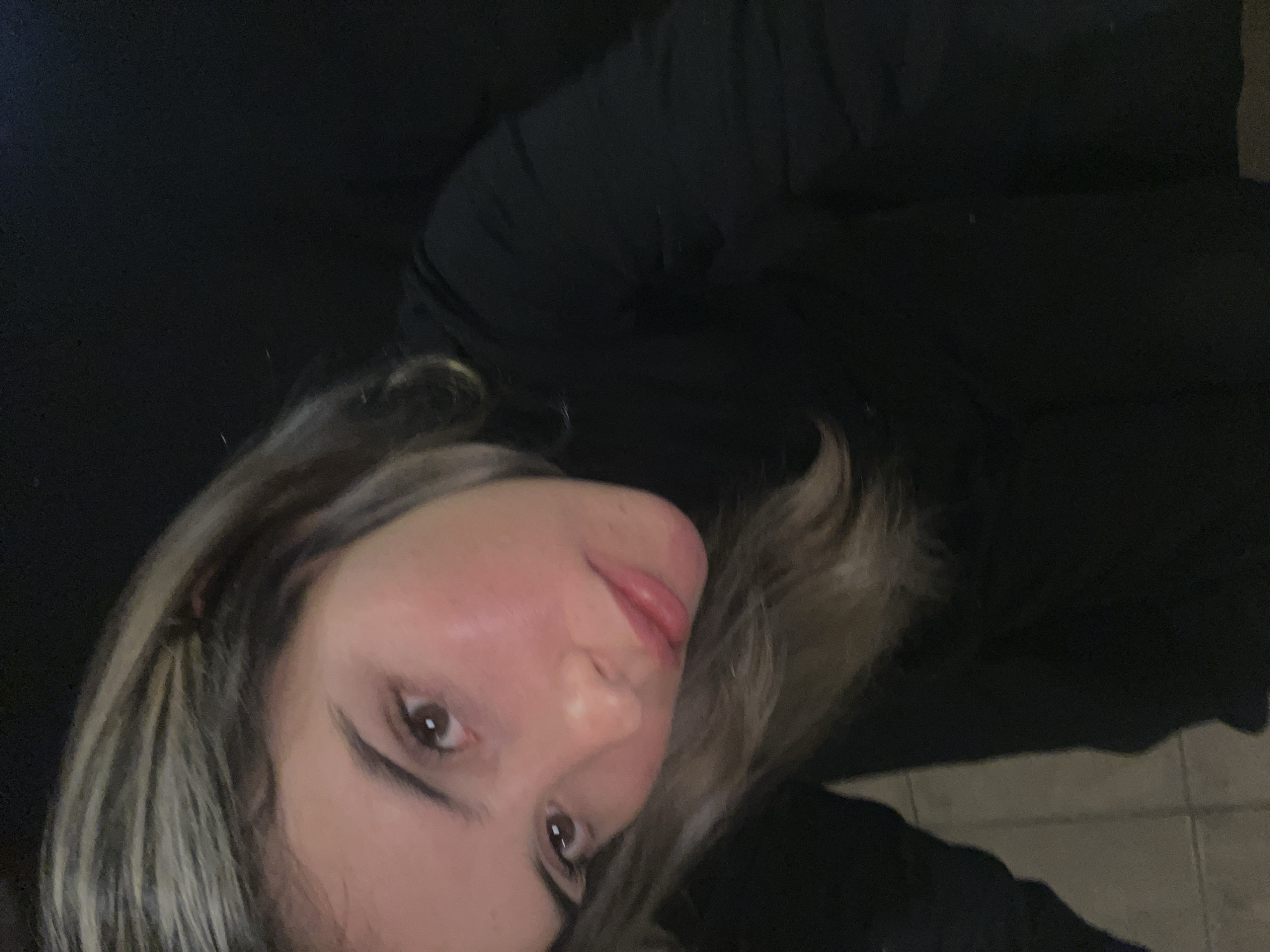 The image shows a close-up of a young woman with blonde-highlighted hair and brown eyes. She is wearing a black turtleneck. Her skin appears flushed, with a hint of pink on her cheeks. The background is dark and out of focus, suggesting an indoor setting. There are no discernible city landmarks or elements that would indicate the location in Sumalao, Argentina.