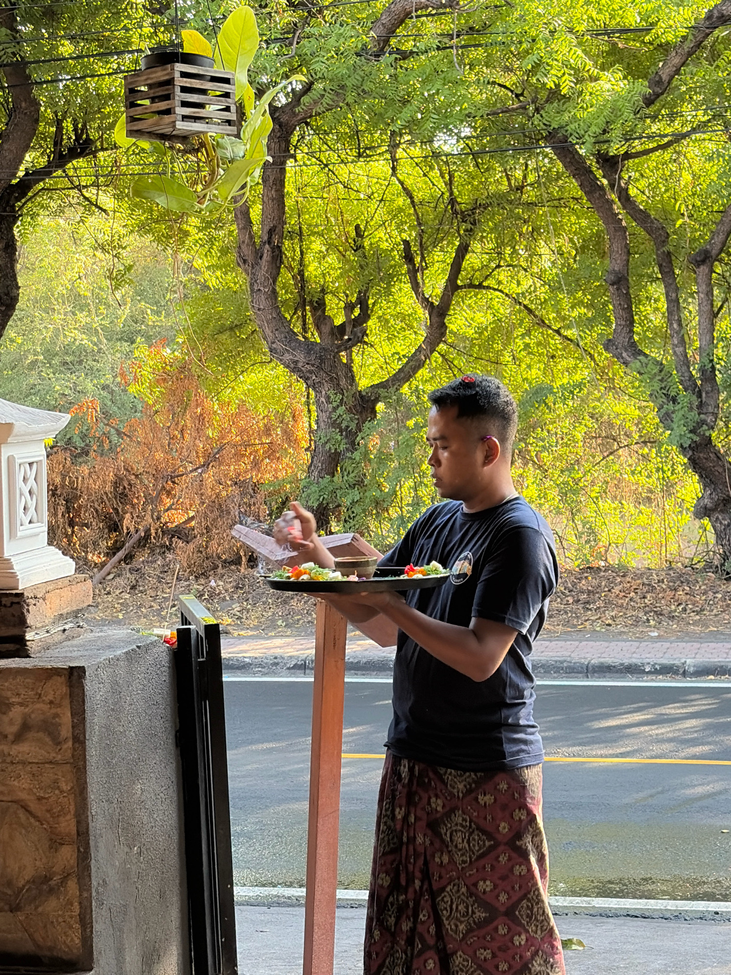A man stands outdoors, holding a tray with offerings. He is wearing a dark t-shirt and a patterned sarong. The tray contains various colorful items, possibly flowers and food. In the background, lush green trees and foliage are visible, with sunlight filtering through the leaves. To the left, a white ornamental structure with intricate carvings is present. The scene appears to be in a tropical environment, possibly a garden or courtyard. The man is positioned near a road, indicated by the asphalt and a yellow line visible in the lower right portion of the image. The overall impression is one of a tranquil outdoor setting, with the man engaged in some form of ritual or presentation.