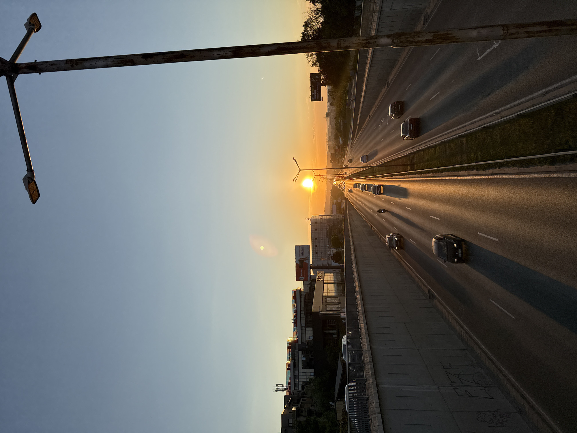 The image depicts a highway scene at sunset in Sofia, Bulgaria. The sun is low on the horizon, casting a warm, golden glow across the asphalt and illuminating the traffic. Several cars and a motorcycle are visible on the multi-lane highway, moving in both directions. The highway is bordered by concrete retaining walls on one side, which show some graffiti. On the other side, a grassy median separates the lanes. In the background, buildings and billboards are visible, suggesting an urban or suburban environment. A tall, weathered lamppost with two lights stands prominently in the foreground on the right side of the frame. The sky is clear, transitioning from a pale blue at the top to a vibrant orange near the horizon.