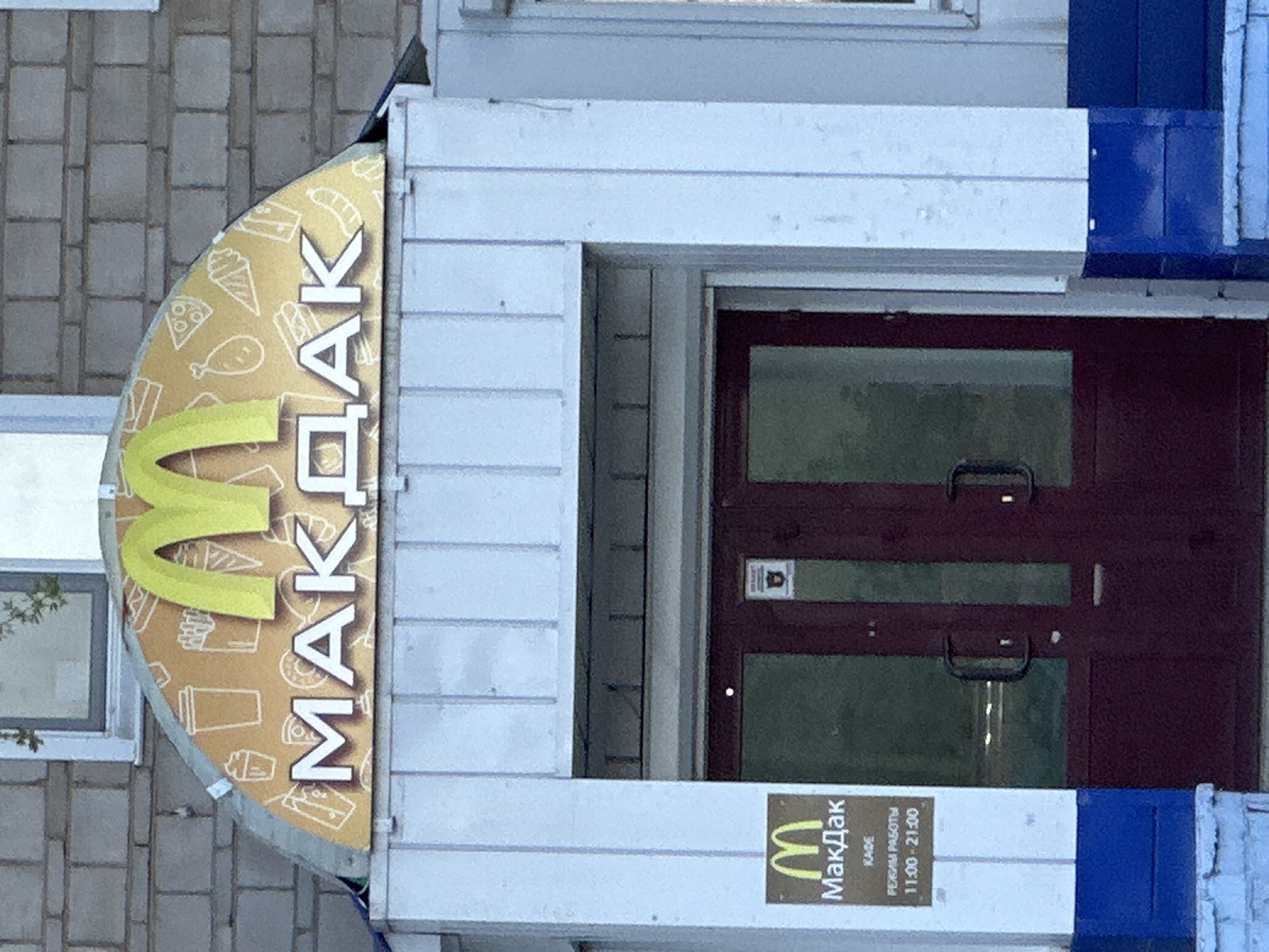 The image depicts a McDonald's restaurant, or rather, a "MakDak" (МакДак), which is a localized brand similar to McDonald's, in Sayansk, Russia. The building is constructed with light-colored siding and blue trim. A half-circle sign with the McDonald's logo and the name "MakDak" in Cyrillic is visible next to the entrance. A smaller sign below the entrance doors also displays the McDonald's logo and the same Cyrillic name, along with the operating hours: 11:00-21:00. The building appears to be a modest structure, not a large, standalone restaurant, possibly a smaller outlet or a café. The environment is urban. The windows of the double door, along with other windows indicate the building is likely more than a small cafe.