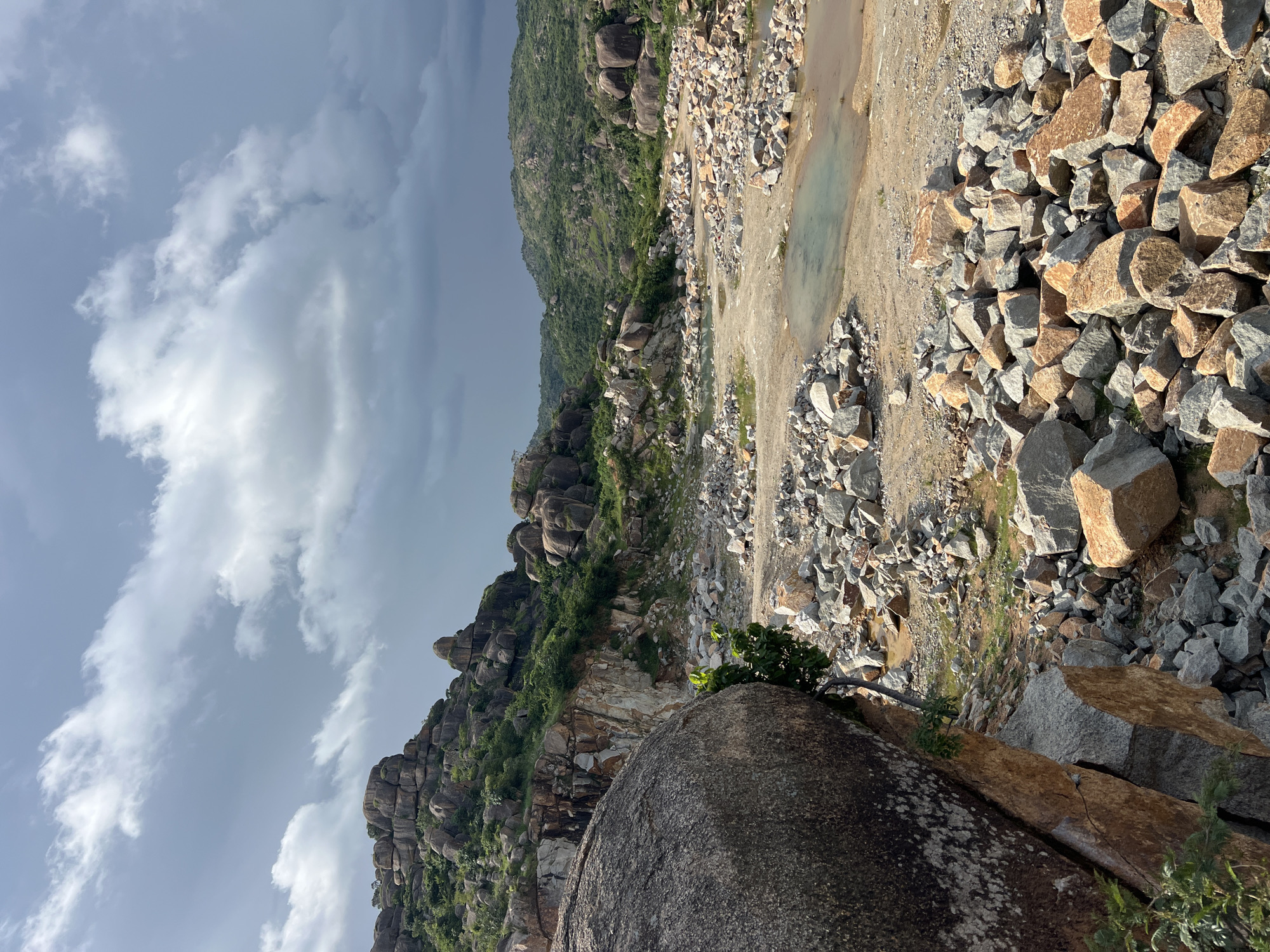 The image depicts a quarry site in Jos, Nigeria, characterized by a rugged, rocky landscape under a dramatic, cloudy sky. In the foreground, a massive, weathered boulder dominates the left side, partially obscuring a steep, rocky embankment. The ground is littered with a vast accumulation of rubble and broken rocks of various sizes and colors, ranging from grey to reddish-brown.
Further into the scene, a shallow body of murky, light blue water is visible, suggesting a small pond or accumulation of rainwater. Beyond this, the terrain rises, revealing more hills and rock formations covered in sparse green vegetation. The distant hills are shrouded in a hazy, subdued light, hinting at either an overcast day or a location experiencing atmospheric dust. The overall impression is one of a raw, industrial landscape shaped by quarrying activities, set against a backdrop of natural, undulating terrain. There are no people or animals visible in the frame.