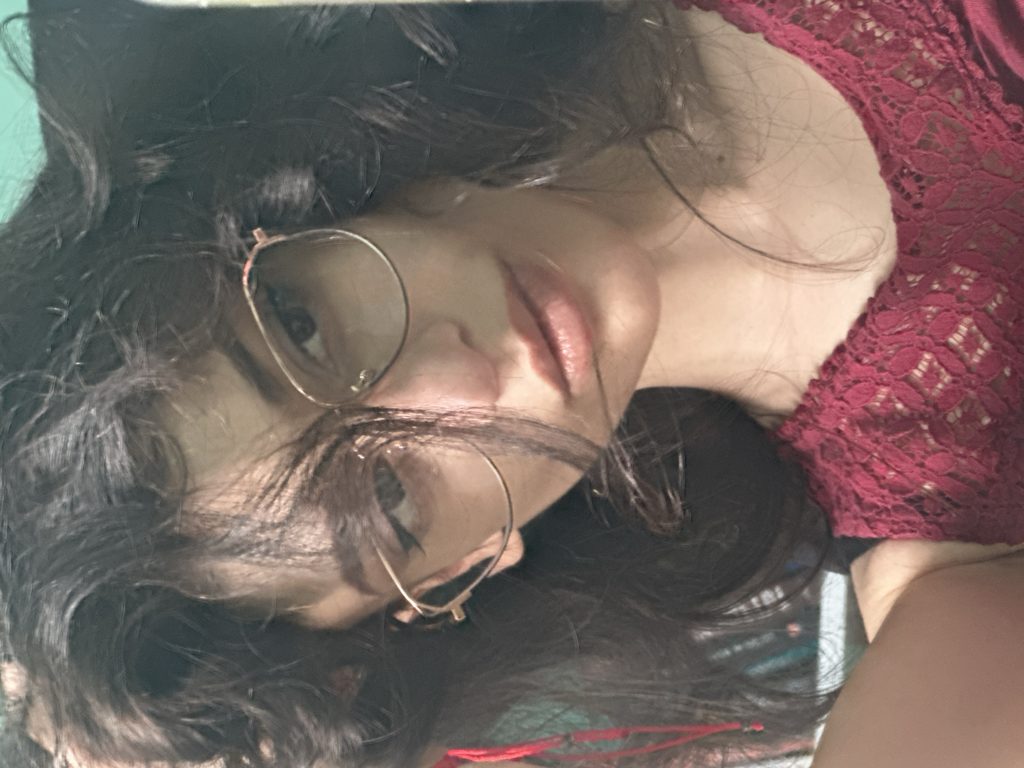 The image is a close-up, selfie-style photograph of a young person with dark, curly hair and glasses. They are wearing a red, lace-trimmed top. The person is looking towards the camera with a neutral expression. The background is out of focus and appears to be an indoor environment with a turquoise wall visible. There are no identifying landmarks or elements in the image that would indicate a specific location within the city of Sofia, Bulgaria.