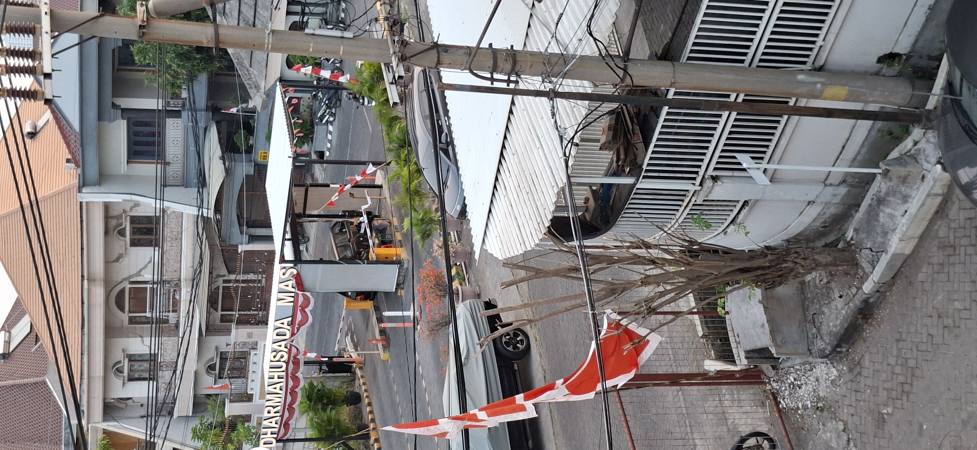 The image shows a street scene in the city of Surabaya, Indonesia, possibly in the vicinity of Dharmahusada Mas. The foreground features a parked silver car, a portion of a truck covered with a tarp, and a large, red and white flag on a pole. Beyond these, an entrance gate is visible with a white awning and the words "DHARMAHUSADA MAS" spelled out in raised, illuminated letters. Several red and white flags are also displayed along the entrance. Two men are seated under the awning, one appearing to be on duty at the gate. A black van is parked at the entrance, and a motorcycle is seen in the background. The architecture of the surrounding buildings is visible, with tiled roofs and balconies. The overall scene depicts a typical street view with elements of commerce and daily life in Indonesia.
