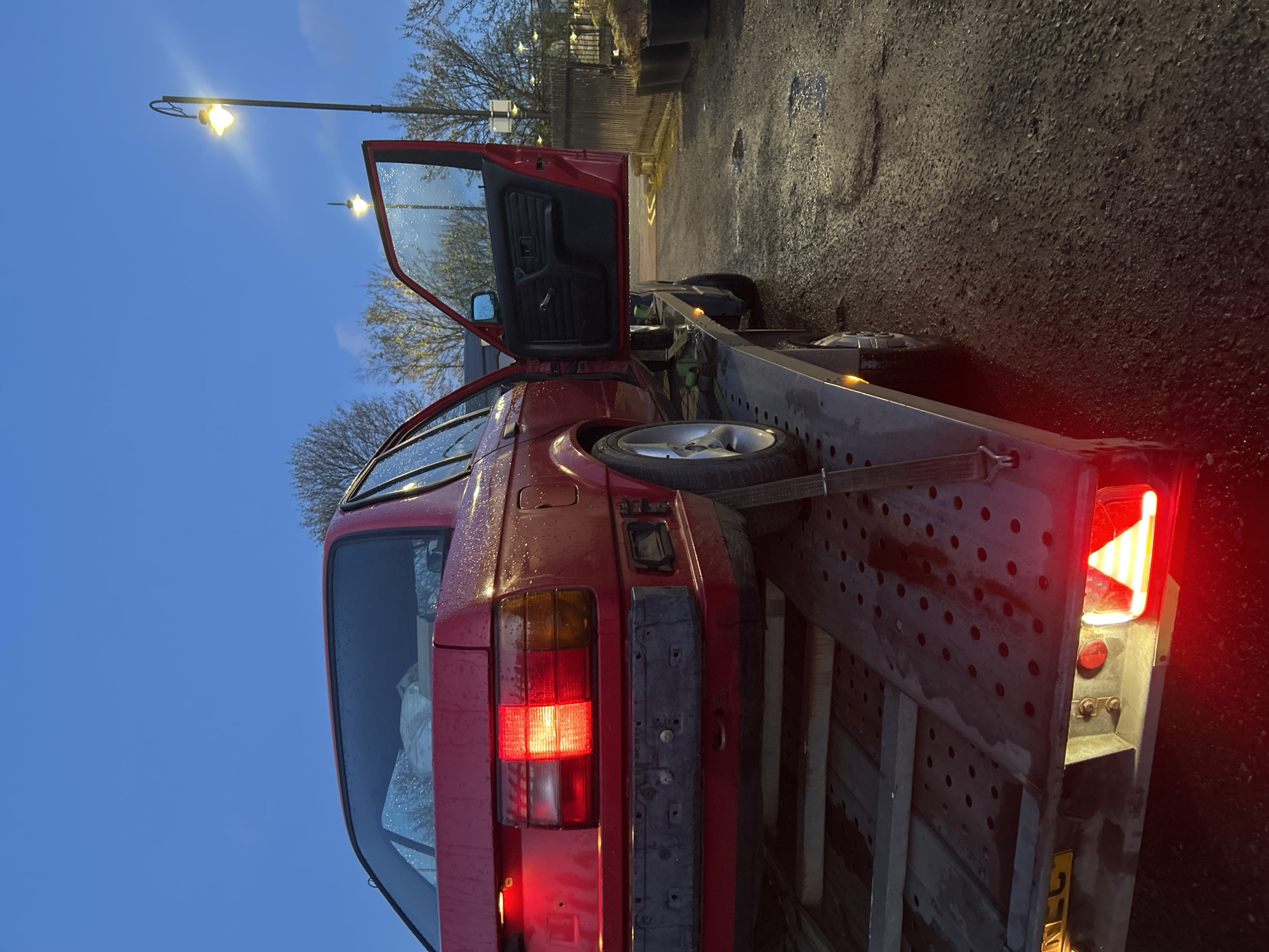 A red car with an open driver's side door is secured to a trailer on a wet asphalt surface. The car is missing its rear bumper. A spare tire is strapped to the trailer near the car's rear. The trailer is parked near a sidewalk adjacent to a fence and some vegetation. A streetlight illuminates the area. The scene appears to be located in an urban setting, potentially in Rutherglen, United Kingdom.
