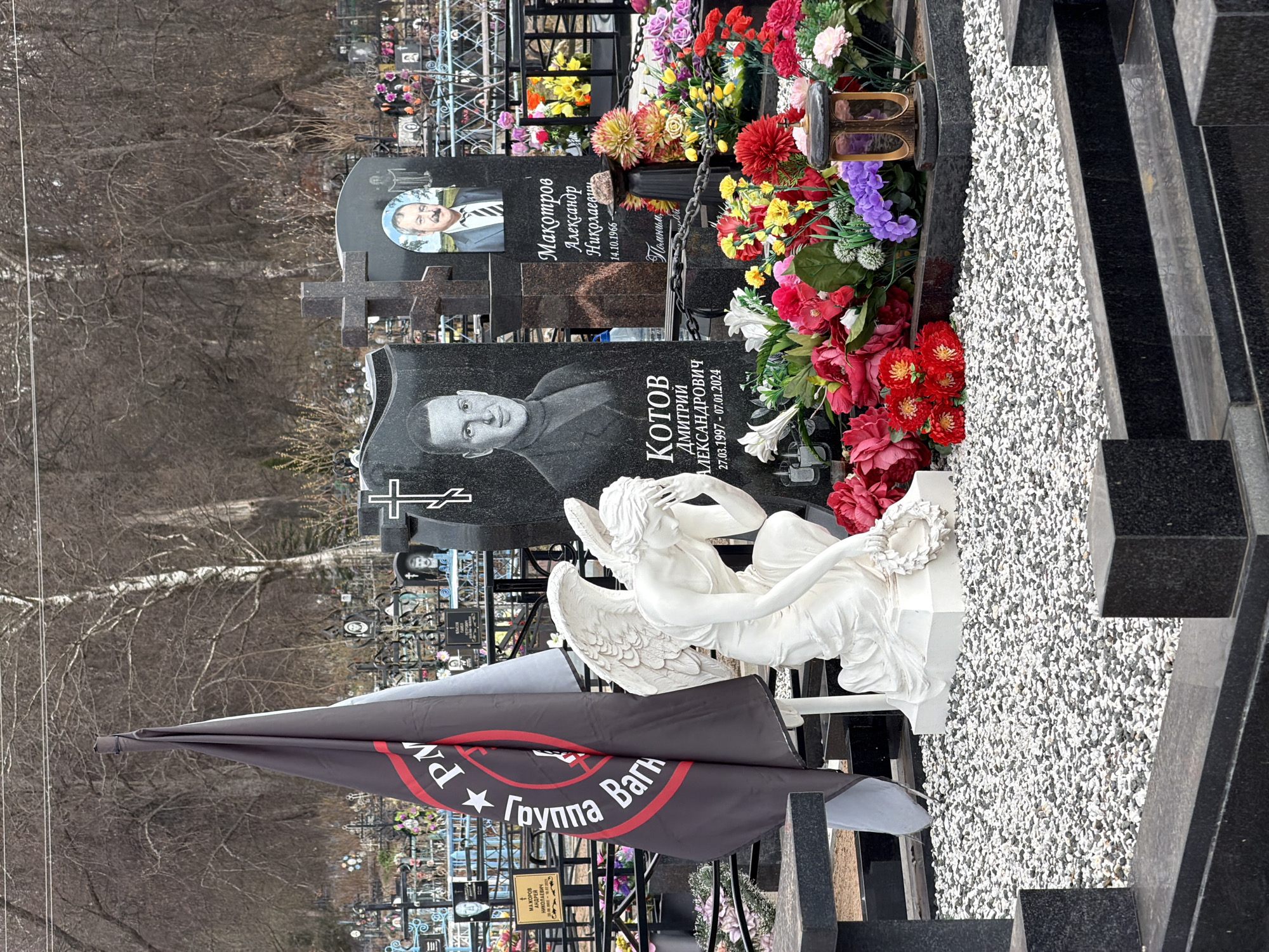 The image depicts a grave site in the city of Бронница, Russia. Two dark granite headstones are visible. One headstone has a photograph of an older man with a mustache. The other headstone has a photograph of a younger man and is inscribed with the name "KOTOB ДМИТРИЙ АЛЕКСАНДРОВИЧ," along with dates "27.03.1997 - 07.01.2024". A white angel statue sits in front of the grave. Red, white, yellow, and purple artificial flowers are arranged near the headstones and statue. A black flag with a red circular border is on display. Text in Cyrillic script on the flag includes the words "ГРУППА ВАГНА" (Wagner Group). The graveyard is located near bare trees and power lines.