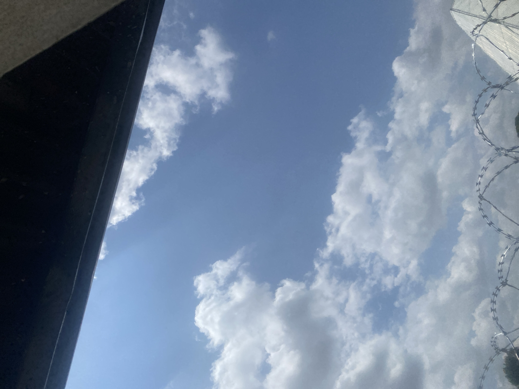 The image shows a clear blue sky with scattered white clouds. In the foreground, there is a dark, horizontal structure across the top of the frame, possibly an overhang or a roofline. Below the sky, a section of razor wire fencing is visible, forming a wavy pattern across the bottom of the image. To the right, a portion of a light-colored, angular structure, possibly a building with a sloped roof or a pyramid-like structure, can be seen behind the razor wire. The overall scene suggests an outdoor environment, possibly a secured area, under a bright sky. There is no indication of the specific location within Lafia, Nigeria.