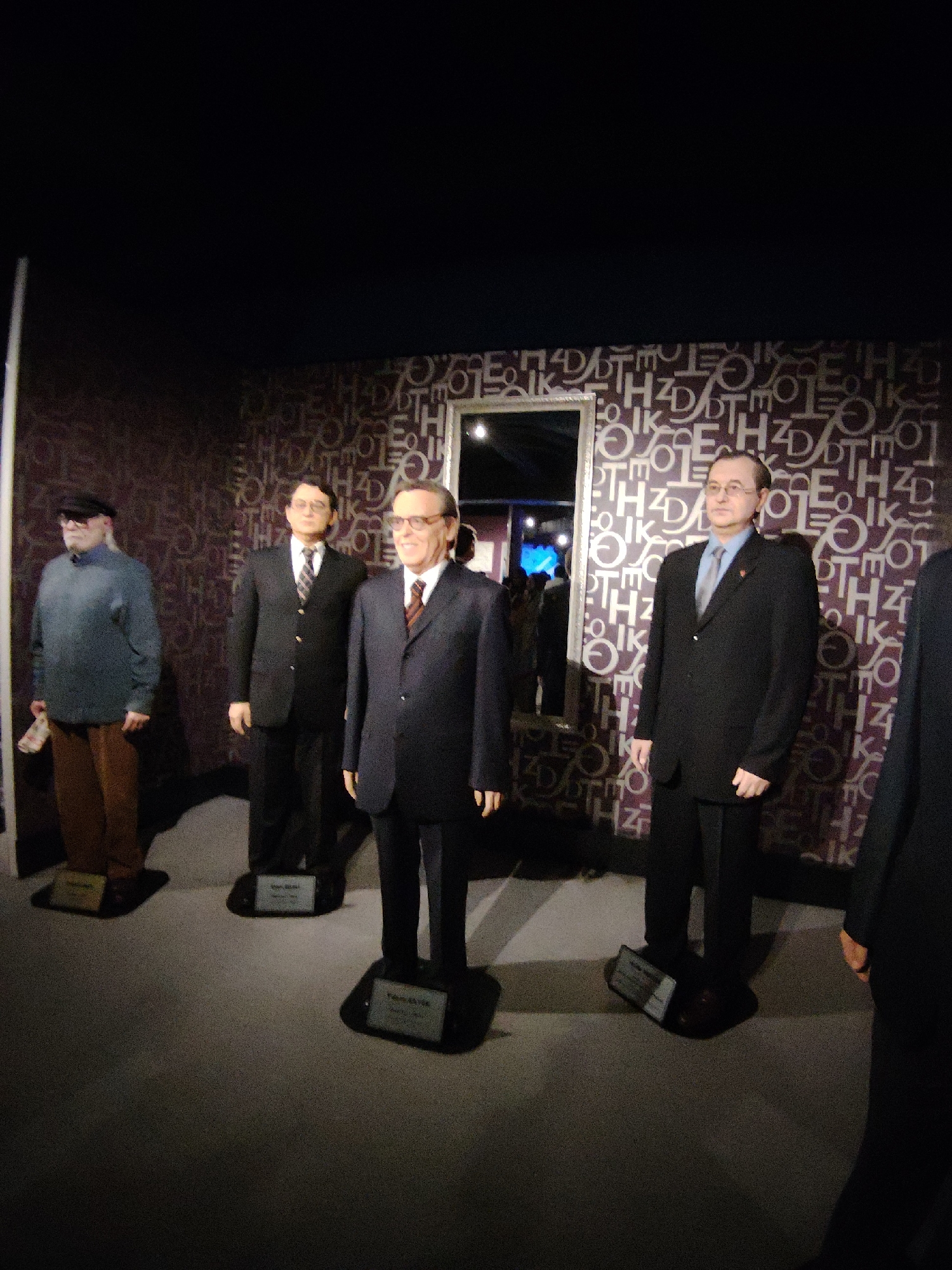 This image captures an indoor scene within a wax museum or exhibition, likely in Eskisehir, Turkey. Four wax figures of prominent Turkish men are displayed, standing on individual dark bases with information plaques in front of them.
From left to right:
1. The leftmost figure is dressed casually in a blue long-sleeved top, brown trousers, and a dark cap. He holds a white object, possibly a newspaper or document, in his left hand. The plaque in front of him reads "Adnan Menderes 1899-1961".
2. The second figure from the left is dressed in a dark suit, white shirt, and a striped tie, wearing glasses. The plaque identifies him as "Vehbi Koç 1901-1996".
3. The third figure from the left, positioned centrally, wears a dark suit, white shirt, and a patterned tie, with glasses. The plaque clearly states "Turgut Özal 1927-1993".
4. The rightmost full figure is dressed in a dark suit, light blue shirt, and a grey tie, also wearing glasses and a small red lapel pin. His plaque is partially obscured but is visible at his feet. A fifth figure, in a dark suit, is partially visible on the far right edge of the frame.
The background features a dark, possibly purple-toned wall covered with a repeating pattern of white or light-colored stylized letters and symbols (e.g., H, D, E, O, Z, K, T). A tall, rectangular mirror with a silver frame is visible on the left side of the background, reflecting light and other parts of the room. The floor is a plain, light grey carpet or surface. The lighting is dim overall, with spotlights illuminating the figures. The scene suggests a dignified presentation of historical or influential personalities.