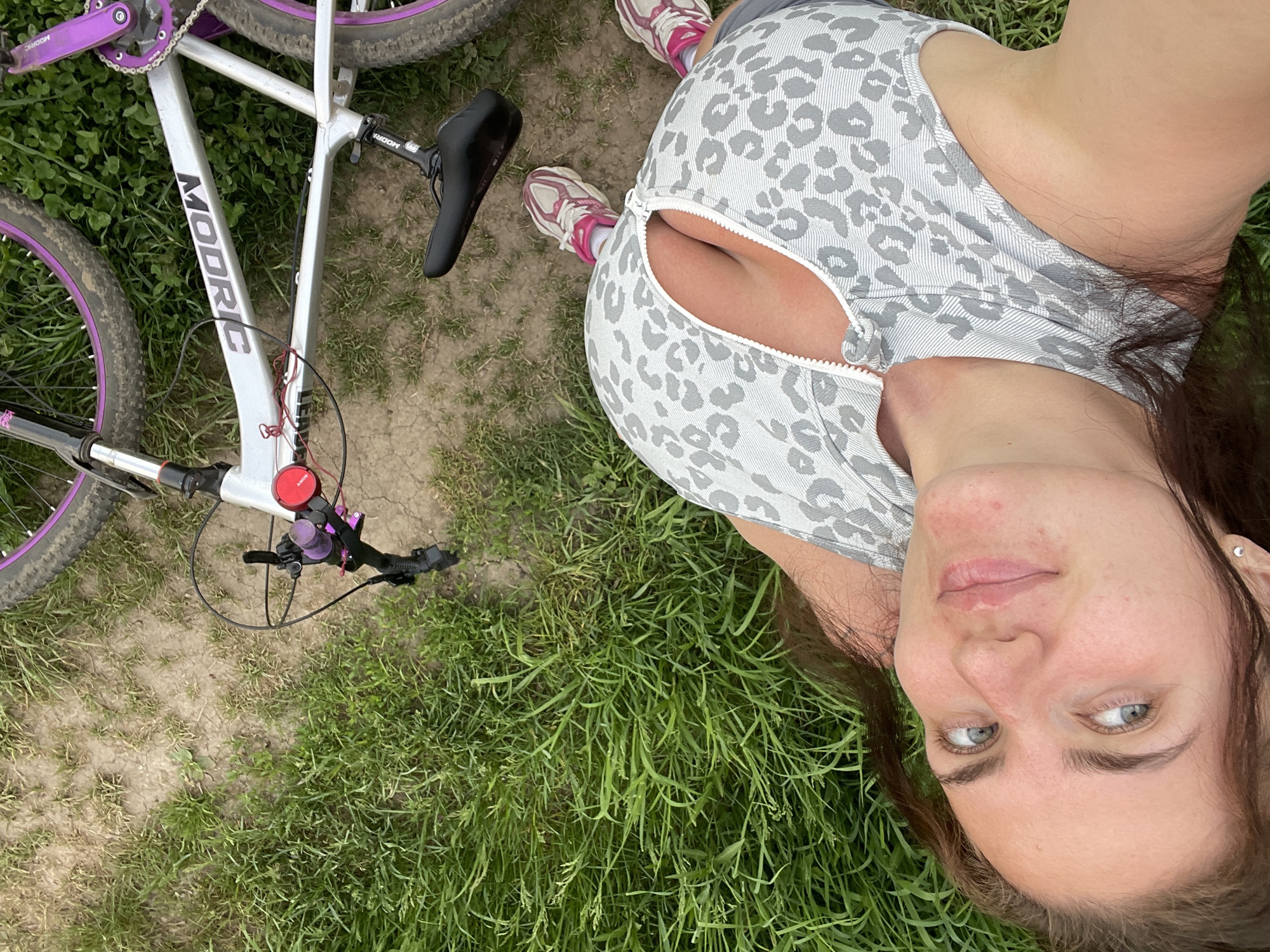 A woman is lying in a field of grass with a white and purple mountain bike nearby. The woman has long brown hair and is wearing a gray and white tank top and pink sneakers. The bike is a Modric brand and is partially visible in the upper left corner of the image. The location is not specified, Ostrov, Russia is not mentioned.