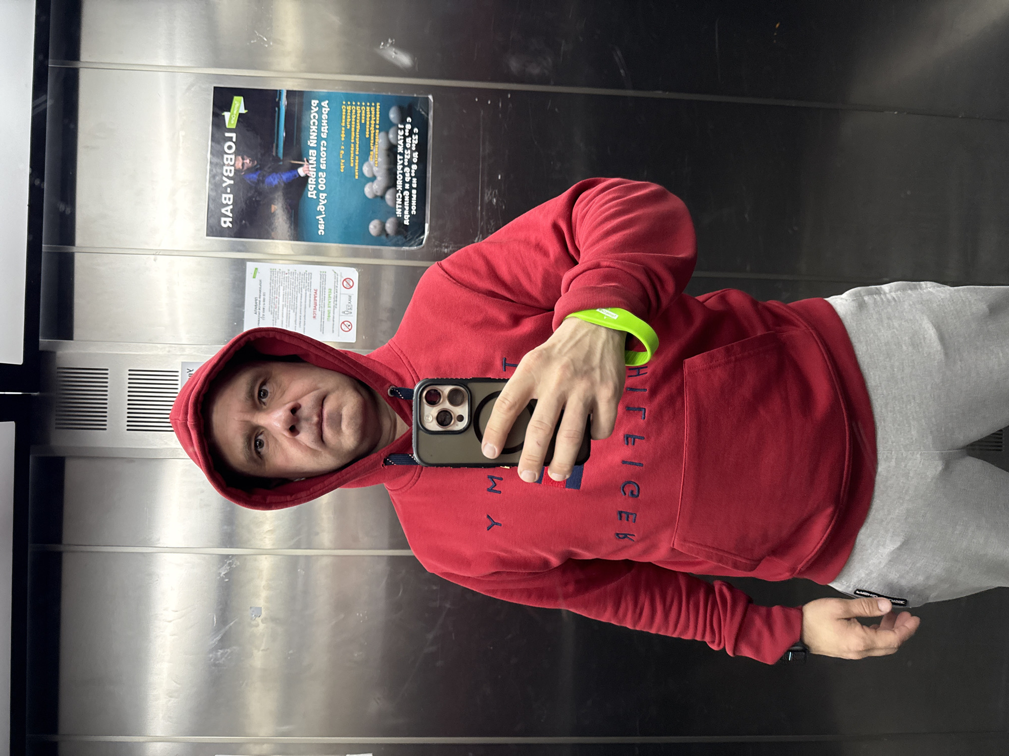 A man in a red Tommy Hilfiger hoodie and grey sweatpants is taking a selfie in an elevator. He is holding a black smartphone with a prominent camera array. The elevator has stainless steel walls, and there are two posters on one wall. One poster advertises "LOBBY-BAR," and the other advertises a pool club, with the text "VUGHTS CLOUS 200 b' Aрас BACCKNN ENUPub." Based on the text and images, the location is in Turgoyak, Russia. The man is wearing a green wristband.