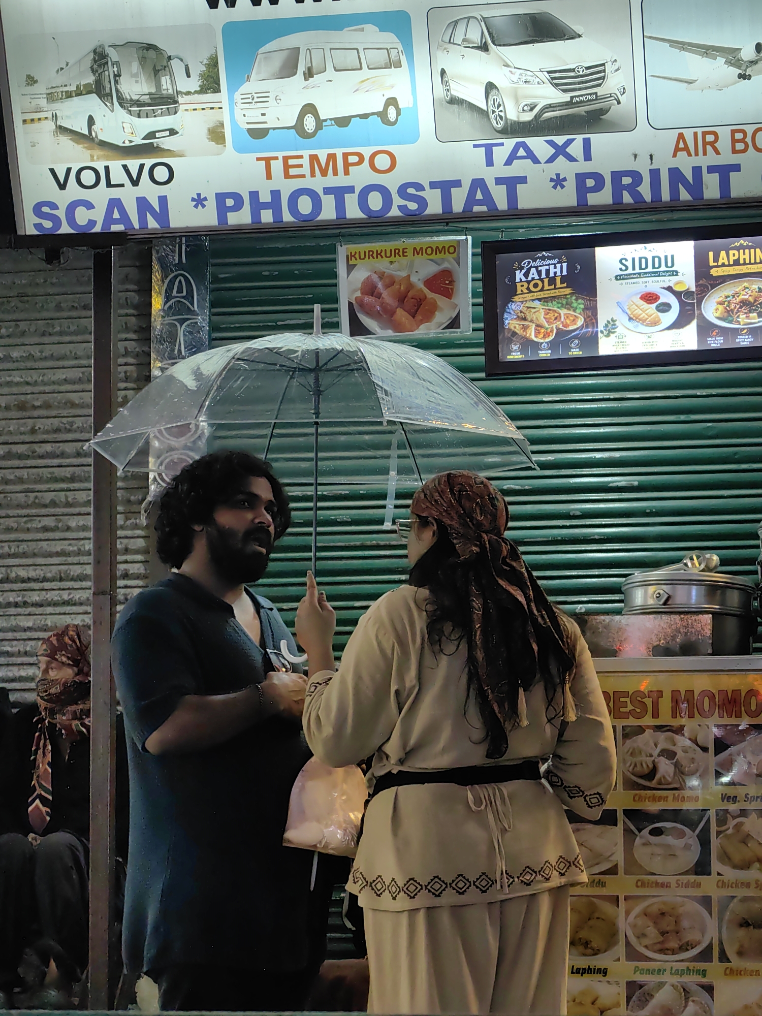 In Manali, India, two individuals stand under a transparent umbrella during a downpour, a common occurrence in this hill station. The backdrop features a vibrant advertisement board for various services, including "VOLVO SCAN * PHOTOSTAT * PRINT," and food stalls displaying pictures of "KURKURE MOMO," "KATHI ROLL," "SIDDU," and "LAPHING." The man, with dark curly hair and a beard, wears a dark t-shirt and points his index finger upwards, possibly to express a thought or question. The woman, with long dark hair adorned with a patterned headscarf, faces away from the camera, dressed in a light-colored outfit. A plastic bag containing what appear to be food items hangs from her hand. The rain-soaked surroundings suggest a busy street scene, typical of Manali's commercial areas.