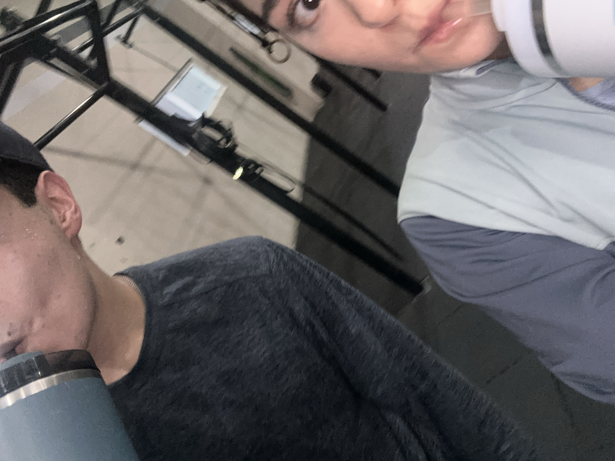 In what appears to be a gym setting, a person on the left is drinking from a light blue hydro flask, with sweat visible on their neck. They are wearing a dark, heathered long-sleeved shirt. To the right, another person is also drinking from a white and gray hydro flask. This individual is wearing a light blue vest over a gray long-sleeved shirt, and sweat is visible on their clothing. The background suggests a gym with metal structures and gym equipment. The overall environment is dimly lit. The image is a close-up, selfie-style shot, focusing on the individuals and their action of drinking.