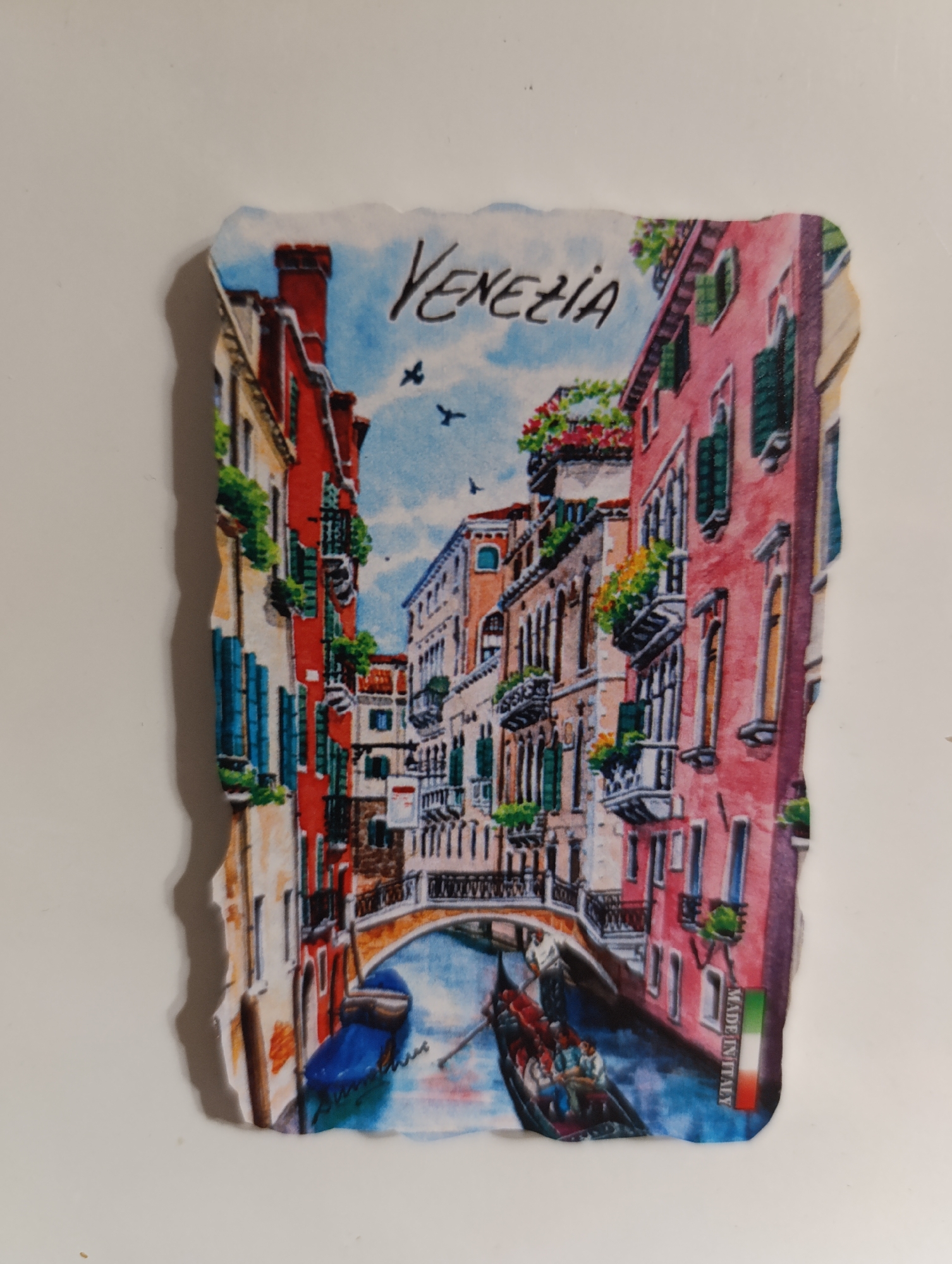 This is a rectangular magnet with a scalloped edge, featuring a watercolor-style depiction of a Venetian canal scene. The buildings on either side are tall and narrow with brightly colored facades, predominantly red and pink. Many windows have green shutters, and some balconies are adorned with potted flowers. A stone bridge arches over the canal in the middle ground. A gondola with several passengers navigates the blue water, and another blue boat is moored to the left. The word "VENEZIA" is written in cursive script across the top of the image. The words "MADE IN ITALY" are printed vertically on the right edge of the magnet, accompanied by the Italian flag. The magnet is set against a plain white background.