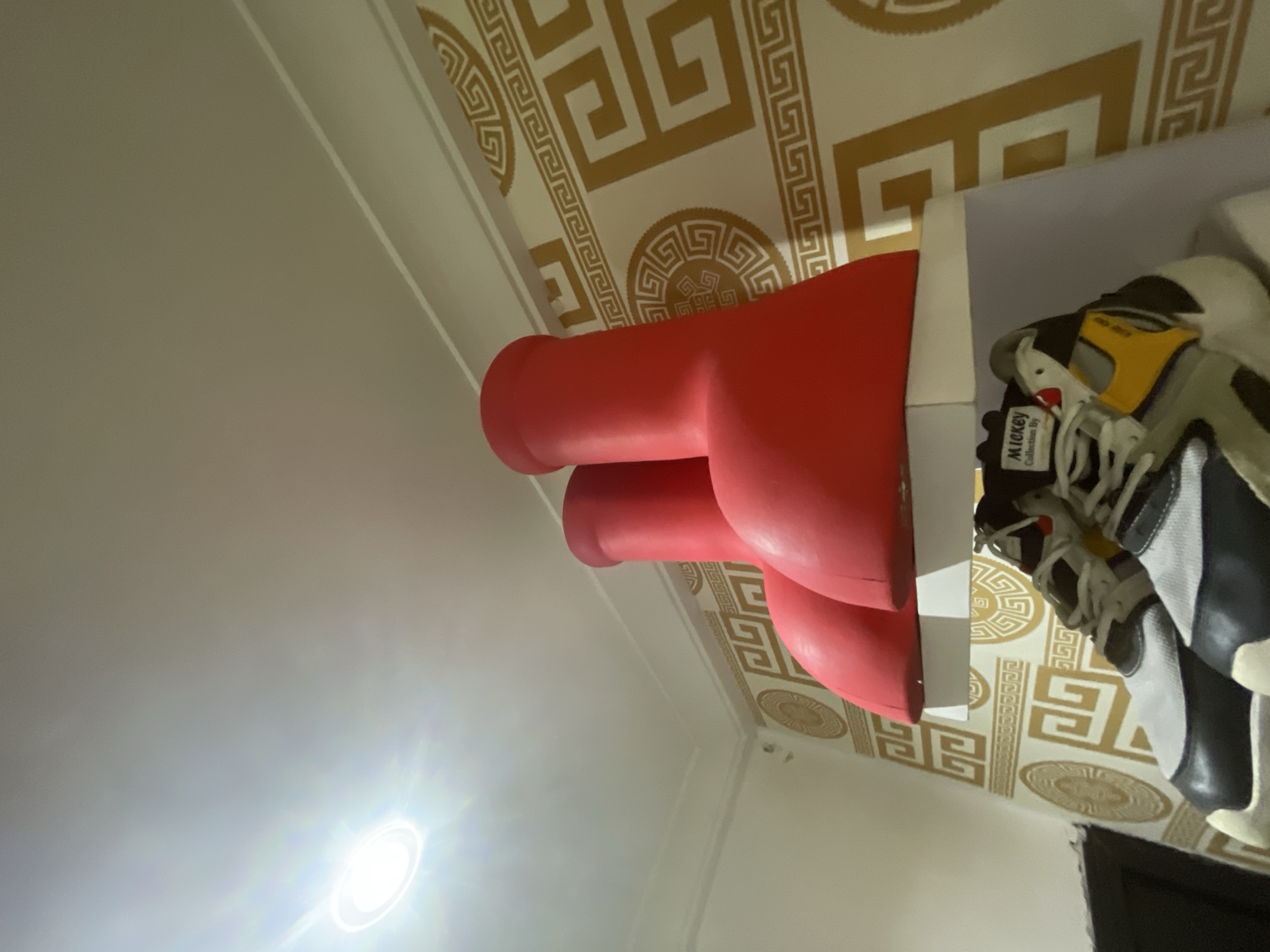 The image, taken in an indoor setting likely in Ado, Nigeria, is captured from a low-angle perspective looking towards the ceiling and a display area. The ceiling is a prominent feature, covered with a wallpaper depicting a decorative Greek key pattern and circular medallions in gold against a light background.
A bright, circular light source, likely a recessed downlight, is active and visible in the upper left portion of the frame, casting a strong glare. On the right side, a large, red, stylized sculpture of a human hand giving a "thumbs up" gesture is displayed on a white shelf or stand. This distinctive red object is positioned next to a pair of athletic shoes or sneakers. The shoes feature a mix of colors including white, black, and yellow. A tag attached to one of the shoes is visible, clearly displaying the text "Mickey Collection By". The walls in the background are light-colored, possibly white or off-white. The scene depicts a static interior display with no human activity taking place.