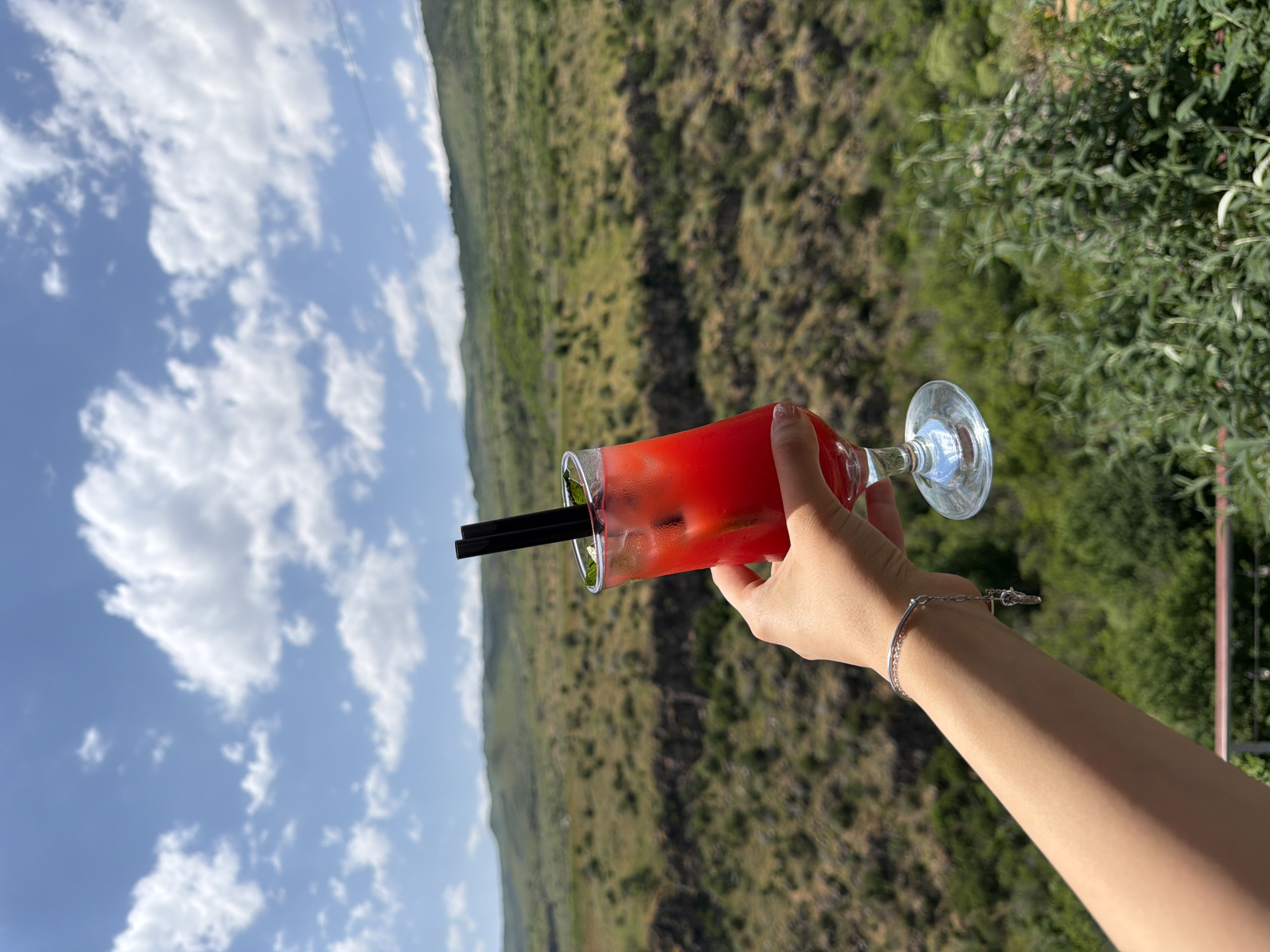 A person's left hand, adorned with a silver bracelet with a charm, holds a tall glass of a vibrant red beverage filled with ice, mint leaves, and two black straws. The drink is set against a backdrop of a lush, green valley with rolling hills and a partly cloudy blue sky. The scene appears to be captured from an elevated vantage point, possibly a restaurant or cafe terrace in Byurakan, Armenia, offering a scenic view of the surrounding natural landscape.