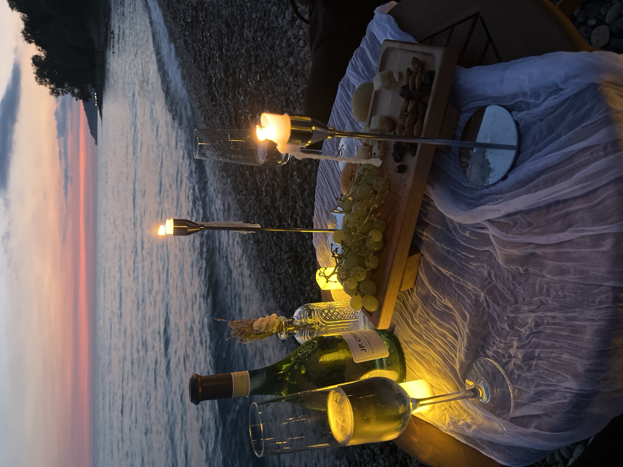 A romantic sunset picnic is set up on a pebble beach along the coast of Sochi, Russia. The scene features a round table covered with a sheer white cloth. On the table, there are two glasses of champagne, one half-full and the other mostly empty, both lit by the warm glow of candles. A green bottle of wine, possibly champagne, stands beside a small decorative vase holding dried flowers. In the center, a wooden serving board holds a bunch of green grapes and two pieces of cheese, while a separate wooden tray contains almonds and raisins. Two candle holders with lit candles are placed strategically, one near the grapes and another towards the right side of the table. The background reveals the tranquil sea meeting a sky painted with hues of orange, pink, and purple from the setting sun. The coastline, lined with trees, curves into the distance. The gently lapping waves and the soft light create a serene and intimate atmosphere.