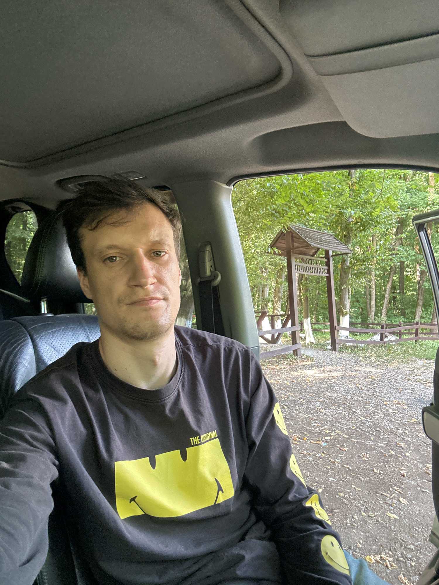 The image shows a man sitting inside a car, taking a selfie. He is wearing a dark long-sleeved shirt with a large yellow smiley face graphic on the front and smaller smiley faces on the sleeves. The car's interior is visible, including the headrest of the seat next to him and a portion of the car's roof. Outside the car, through the open door and windshield, a wooden signpost with Cyrillic text is visible in a wooded area. The ground outside is covered in gravel and fallen leaves. The setting appears to be a natural or park-like environment, possibly near the village of Shatava, Ukraine, given the signage in Cyrillic script. The man has short, dark hair and is looking directly at the camera with a neutral expression. The overall lighting suggests it is daytime.