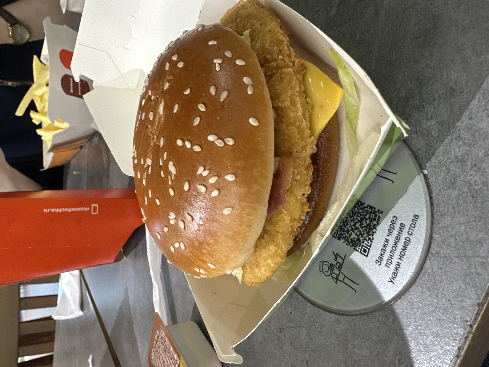 A partially eaten chicken burger sits in its cardboard container on a gray tabletop, alongside a small container of fries and a red box labeled "vkusnoitochka.ru." The burger bun is topped with sesame seeds, and the contents include a fried chicken fillet, lettuce, cheese, and bacon. A coaster is also visible, featuring a QR code and Russian text that translates to "Order via the application. Indicate the table number." The scene suggests a fast-food meal at a Vkusno i tochka restaurant, likely in Kazan, Russia, based on the restaurant's branding and the Russian language used on the coaster. A partial view of a person and their watch is visible in the background, indicating they are seated at the table.