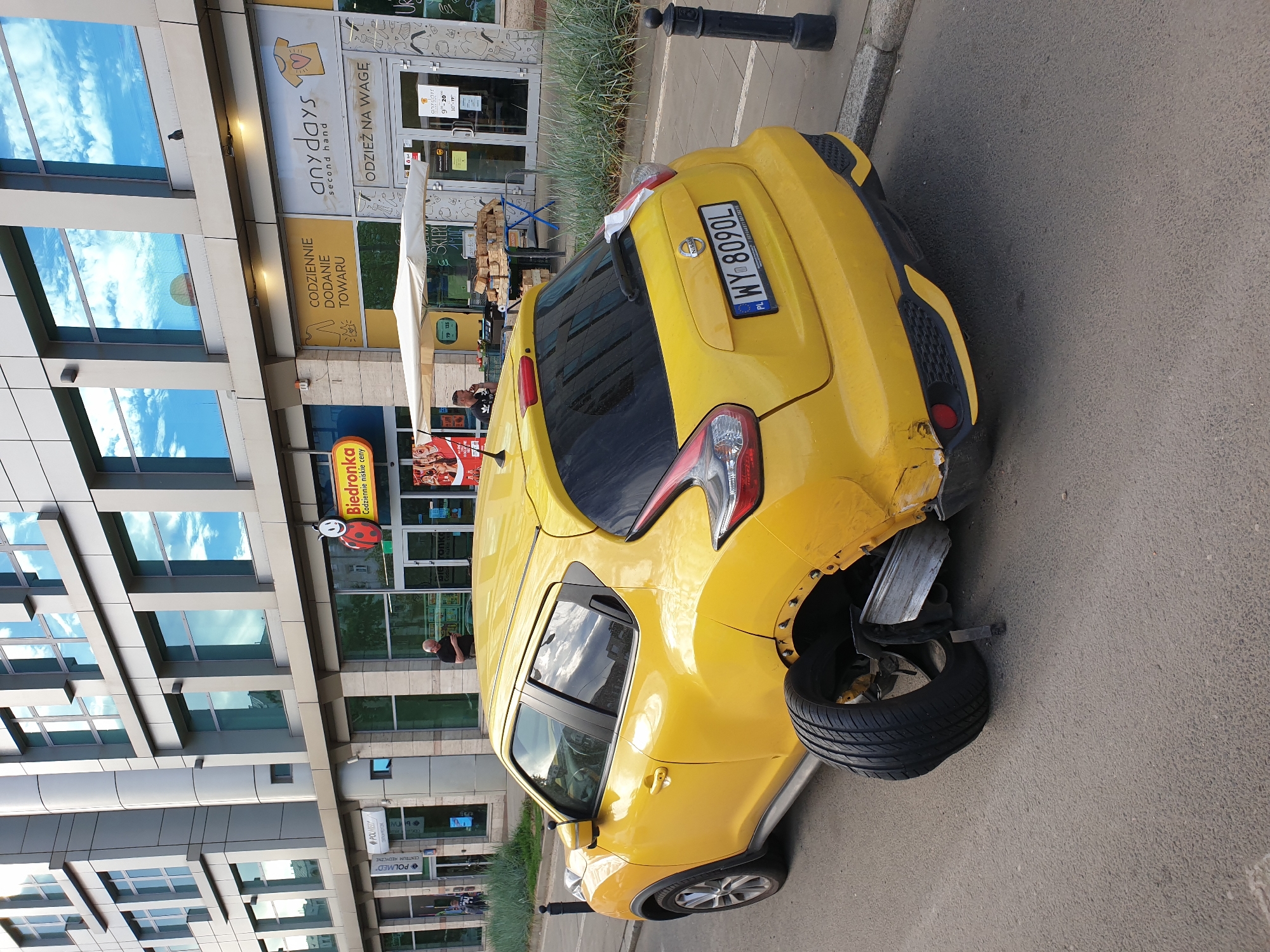 The image depicts a yellow Nissan Juke parked on a street in Warsaw, Poland. The car has sustained significant damage to its rear right side, with the wheel detached and lying on the pavement. The bodywork is crumpled and torn around the wheel well, and a piece of metal bracing is exposed.
In the background, a modern building with large glass windows reflects a cloudy sky. Various shop signs are visible on the building's facade. One sign reads "anydays second hand," another "CODZIENNIE DODANE TOWARU," and a red and yellow circular sign features a ladybug and the text "Biedronka" with "Codziennie niskie ceny" below it.
A few people are present. One person, wearing a black t-shirt, stands near a shop entrance, seemingly observing the damaged car or the surroundings. Another person is partially visible through the shop windows.
The scene appears to be a street in a commercial area, likely during daytime. The lighting suggests it is not early morning or late evening. The overall impression is one of an unfortunate incident, with the damaged vehicle being the central focus against the backdrop of a busy urban environment. The image is oriented vertically.
