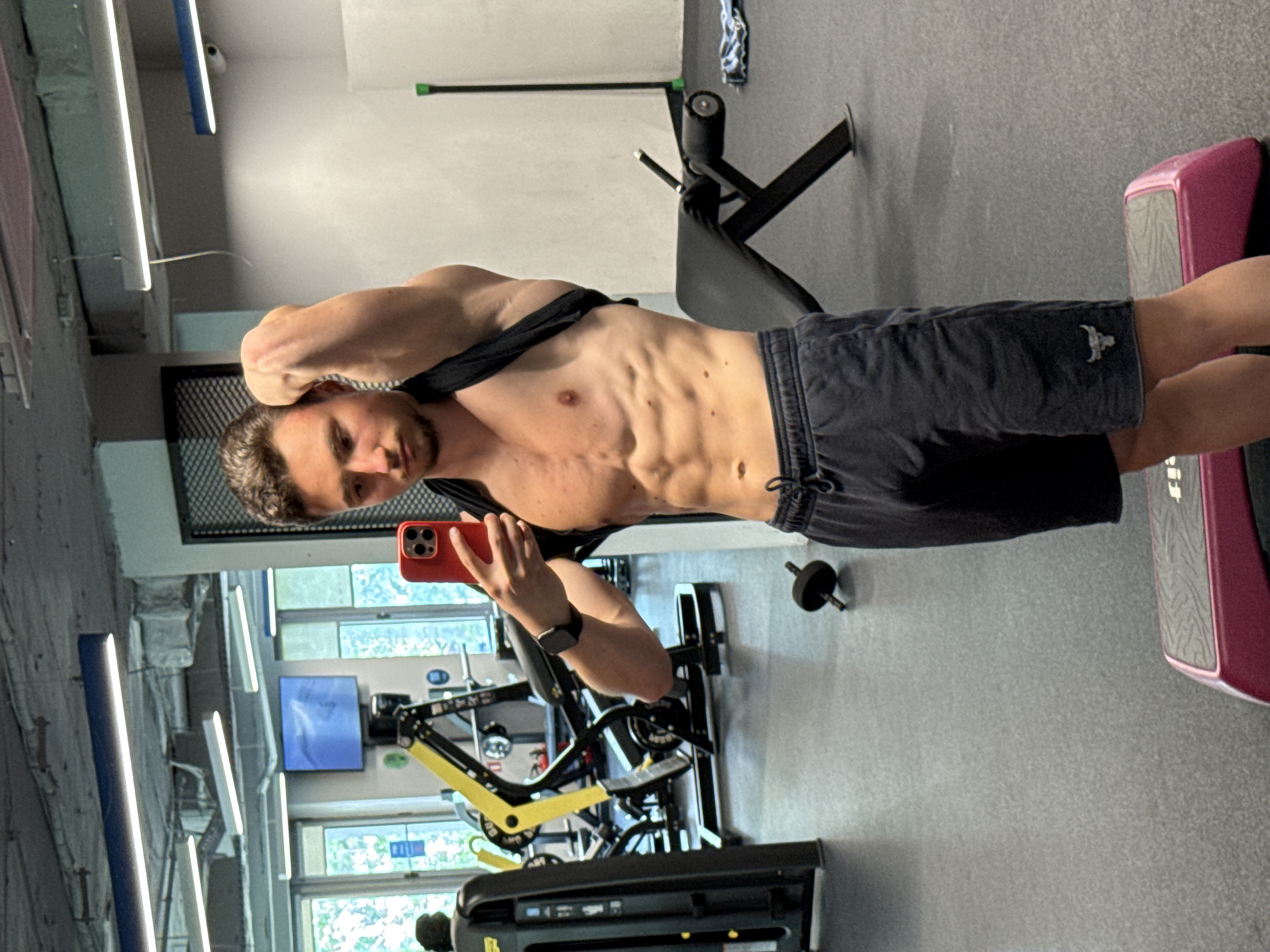 A man is taking a selfie in a gym. He is shirtless, wearing dark shorts, and holding a red smartphone in his right hand. He is standing in front of a mirror, posing to show his physique. Behind him are various pieces of gym equipment, including weight machines, exercise benches, and steps. The gym appears to be well-lit and modern. The shorts have a bull's head logo. The gym overlooks trees. The location is most likely in Sofia, Bulgaria.