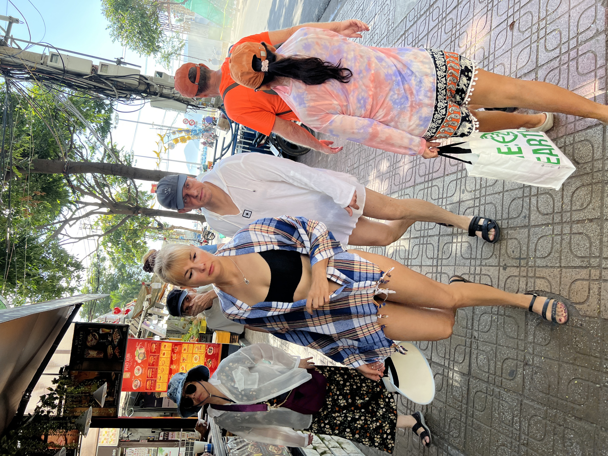 In Nha Trang, Vietnam, a group of four individuals walks along a sidewalk lined with shops and trees. The foreground features a woman in a blue and white plaid shirt worn open over a black bikini top, denim shorts, and black sandals. She carries a white, flat disc-shaped object. To her left, a woman wearing a wide-brimmed hat, a sheer white jacket, and a floral skirt walks with a purple crossbody bag. Behind the plaid-shirted woman, a man in a white hooded tunic and shorts walks with black sandals. Further back, a person in an orange shirt and an orange cap is followed by a woman in a tie-dye shirt and patterned shorts, holding a white reusable shopping bag with green lettering that reads "RE EARTH." The sidewalk is paved with patterned tiles, and a street with parked motorcycles is visible to the right. The background shows buildings, electrical wires, trees, and a glimpse of the sea under a bright, sunny sky. The scene appears to be a casual stroll in a bustling urban environment.