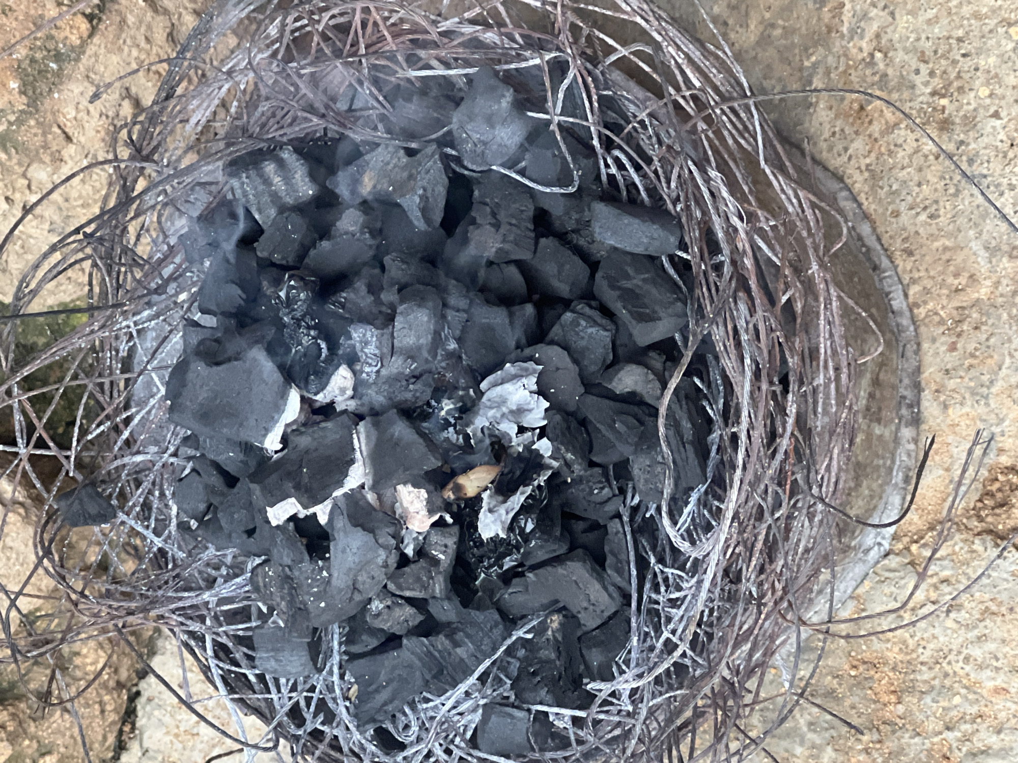 The image shows a pile of charcoal in a nest-like structure made of wire. The charcoal is mostly black and appears to be broken into irregular pieces. Some of the charcoal pieces have white or pinkish ash on them. There are also wisps of smoke rising from the charcoal, indicating that it may still be hot or smoldering. The wire structure is made of thin, intertwined wires, some of which are rusted. The wire structure appears to be resting on a rough, stony surface, possibly the ground or a paved area. The background is out of focus but appears to be a rough, rocky or earthy texture. The scene is photographed from a high angle, looking down into the pile of charcoal. The image has a naturalistic lighting. The context of Jos, Nigeria, suggests this could be a scene related to cooking, heating, or artisanal activities in the city.