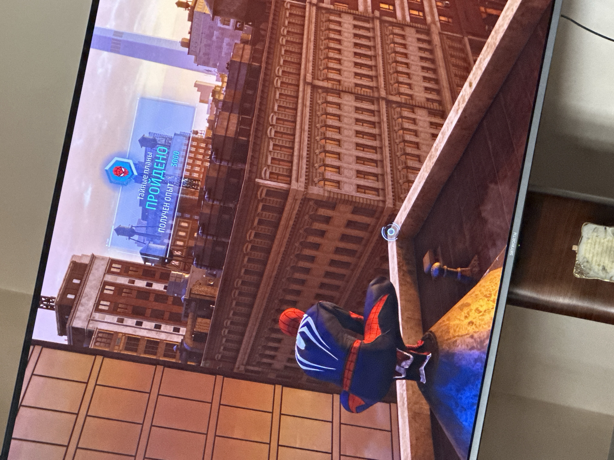 A television screen displays a scene from a video game, featuring a character resembling Spider-Man perched on the edge of a building in a city environment, which appears to be New York City. The character is wearing a blue and red suit with white markings. He is positioned on a ledge against the backdrop of tall, older-style buildings with multiple windows. A taller, modern skyscraper is visible in the distance. An overlay on the screen indicates "Secret Plans" and the completion of an objective, awarding the player 3000 experience points.
