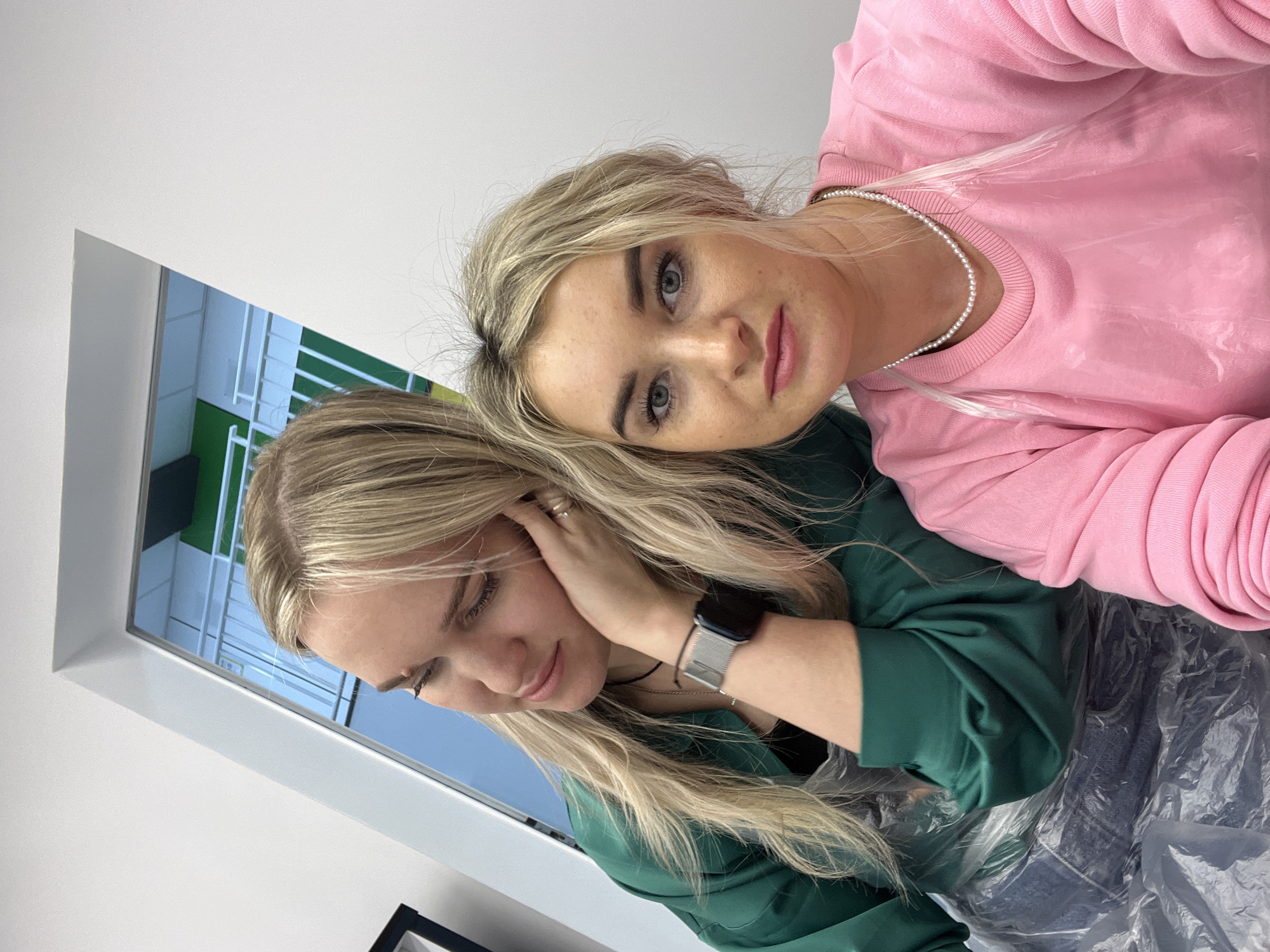 Two young women are in what appears to be a dental clinic or medical office. One woman wears a pink sweatshirt and a pearl necklace. The other wears a green long-sleeved shirt. The woman in green is touching her hair near her ear and has a wristwatch on her left wrist. A framed image of a building that looks like it may be the Izhevsk State Medical Academy (ISMA) is visible. They seem to be sitting in a dental chair. There are clear plastic covers over parts of the dental chair.
