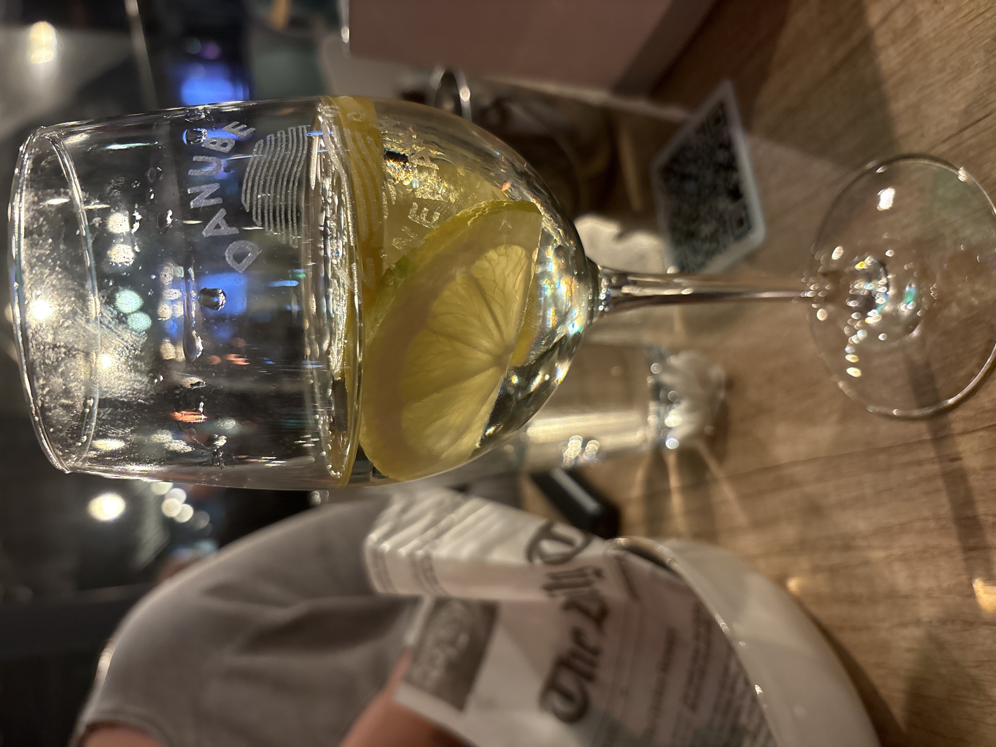 The image shows a clear glass with the inscription "DANUBE" near the rim, containing water and lemon slices. The glass is placed on a table, next to a newspaper or printed material titled "The City". The scene appears to be indoors, with a blurred background suggesting an urban setting. Given the inscription "DANUBE" and the presence of "The City" newspaper, it is plausible to assume this scene is in Sofia, Bulgaria, where the Danube river is a significant geographical landmark and urban newspapers exist. There's a person in a gray shirt visible partially in the foreground. A QR code and other indistinct objects are also in the background.