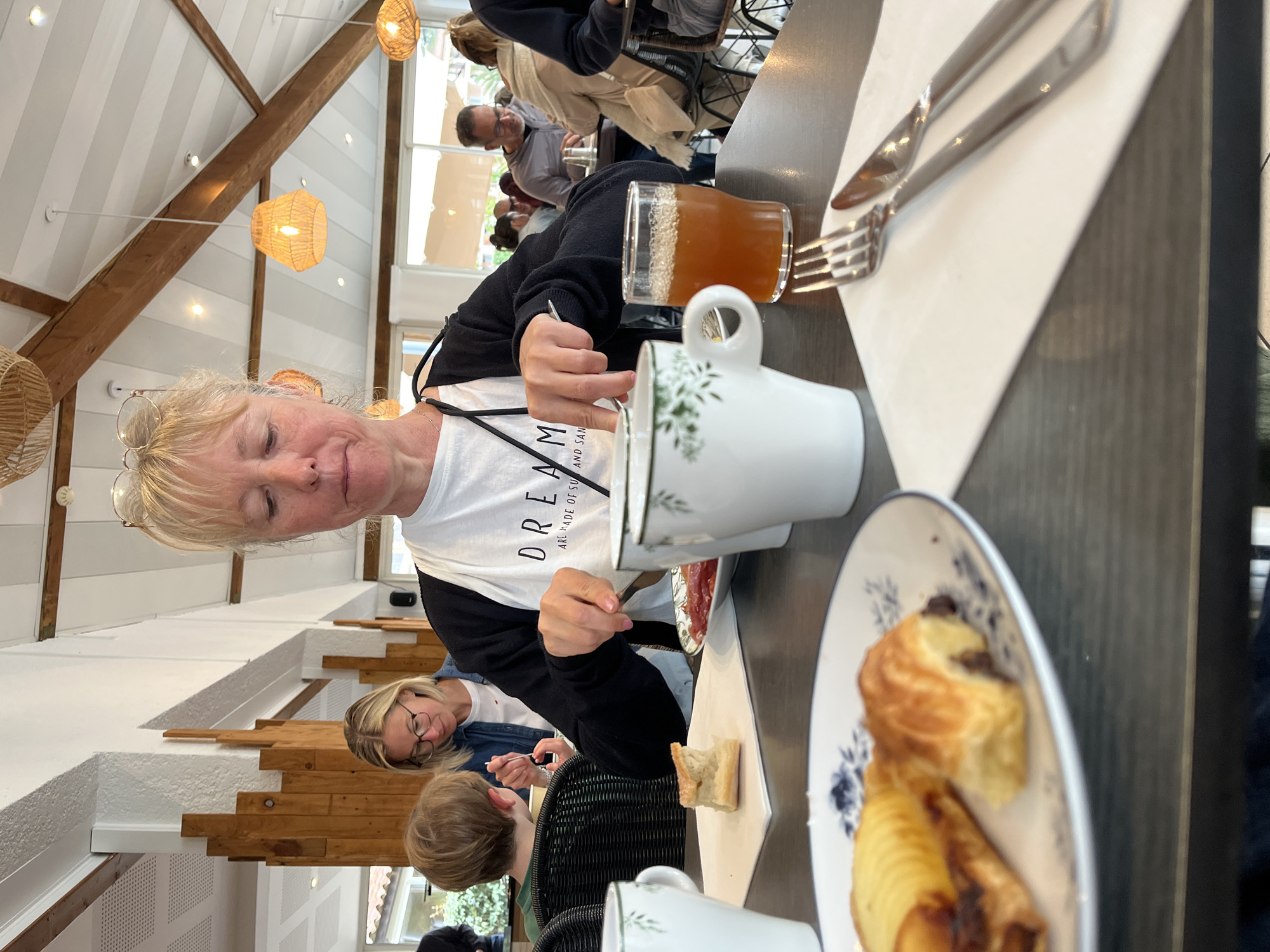 Scene: A group of people are seated at a table in a bright, indoor dining area.
People: An older woman with blonde hair is seated at the table and looking down. A younger woman wearing glasses and a denim jacket is visible to the left of the older woman. The top of the head of a child is also visible at the table. Other patrons are seated at tables in the background.
Objects: A white mug with a green floral design, a glass of amber-colored liquid, a knife and fork on a white napkin, and a pastry on a small plate with blue designs are visible on the table. Other pastries, bread, and a plate of meat are also on the table.
Environment: The indoor dining area has white walls and a wooden beam ceiling with pendant lighting. The location is in the city of Grimaud, France.
Actions: The older woman appears to be stirring something in her mug.
Interactions: People are eating and interacting with one another at the table.