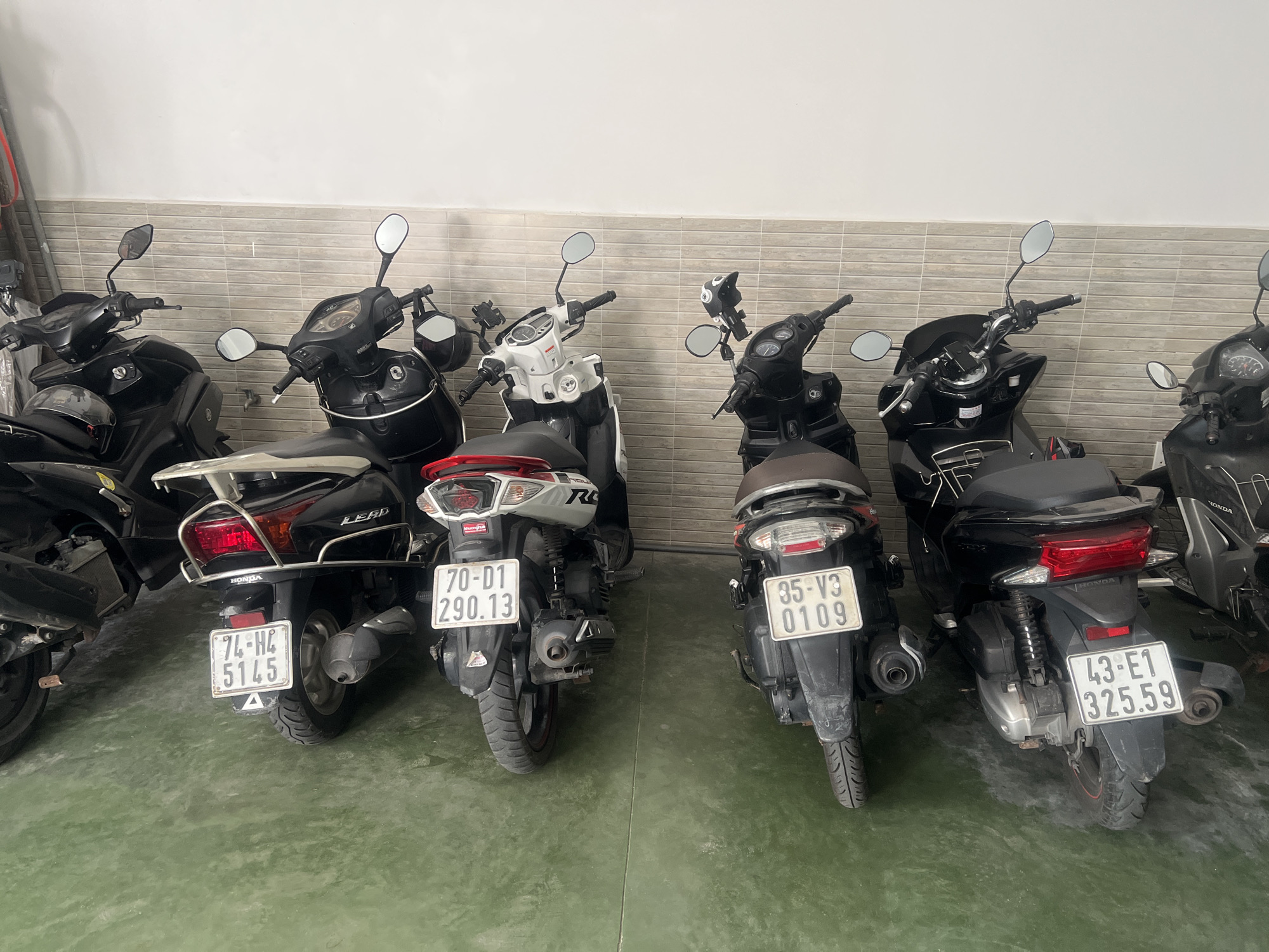 The image shows a row of parked motorbikes in what appears to be a covered parking area, possibly an underground garage or a covered lot. The floor is a speckled green concrete, and the wall behind the motorbikes is tiled with rectangular beige tiles. There are at least six motorbikes visible, all facing away from the camera. Some of the motorbikes have license plates, and the license plate details indicate they are registered in Vietnam, with the visible plates showing combinations of numbers and Vietnamese characters, and some plates bearing the number "43" which signifies registration in Đà Nẵng city. The lighting is somewhat dim, suggesting an indoor or shaded environment. The motorbikes are of various models and colors, mostly black, with one white and black model visible. None of the motorbikes appear to be in motion, and no people are present in the image.