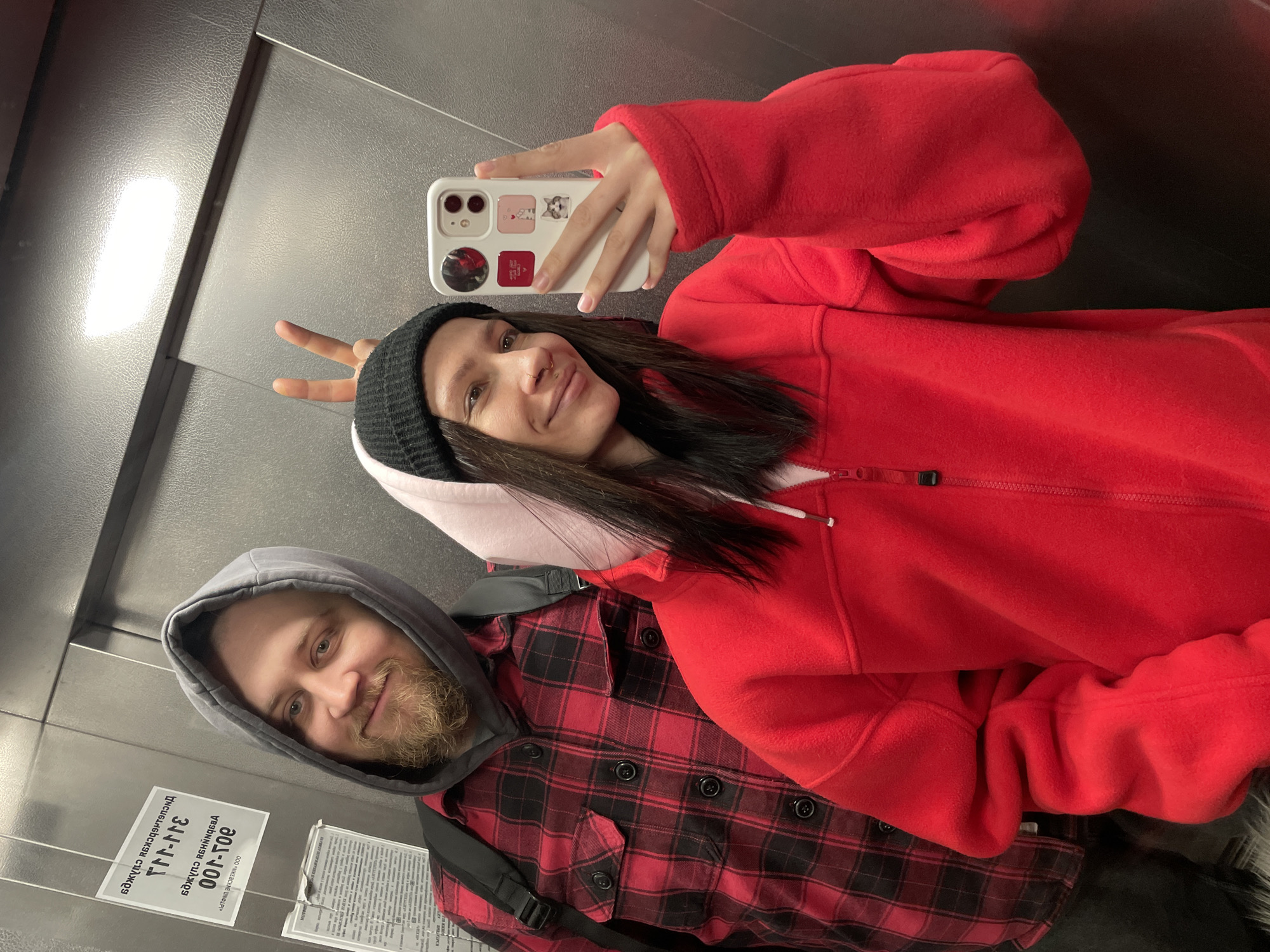 A photograph, rotated counter-clockwise, shows two individuals in what appears to be an elevator in Izhevsk, Russia. The woman, wearing a red hooded fleece jacket and a black beanie, holds up a smartphone with a white case decorated with images, possibly stickers or decorative elements. She makes a “peace” or “victory” sign with her fingers. Next to her, the man, wearing a grey hooded sweatshirt and a red and black plaid button-down shirt, has a beard and is smiling. A black strap, probably from a bag, is visible across his torso. On the elevator wall behind them, there are two notices with Cyrillic text, indicating that the location is in Izhevsk, Russia. The elevator's interior is a grey metal material with a subtle texture.