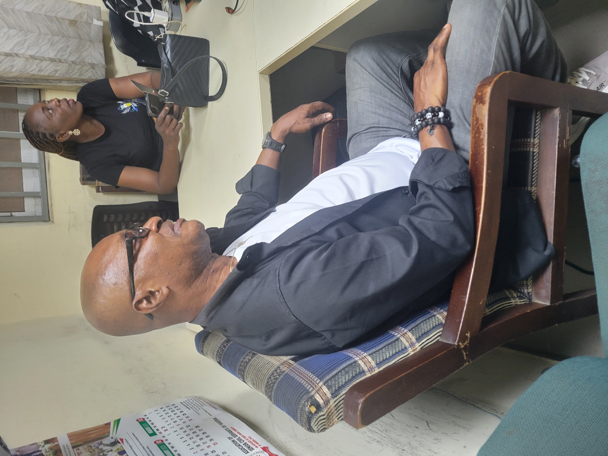 A man is sitting in a wooden chair in what appears to be an office setting. The man is bald and wearing glasses, a white shirt, and a black jacket. He has a bracelet on his wrist and appears to be looking up and to the left. The chair has a cushion with a blue and brown plaid pattern. Another woman is visible, sitting at a table, holding a phone and a handbag. A calendar is visible in the lower left corner, with the text "ASSOCIATION OF SENIOR CIVIL SERVANTS OF NIGERIA", indicating that this is likely an office associated with that organization. Given the location of the organization in Nigeria, it is possible that this is located in the city of Abuja, Nigeria.
