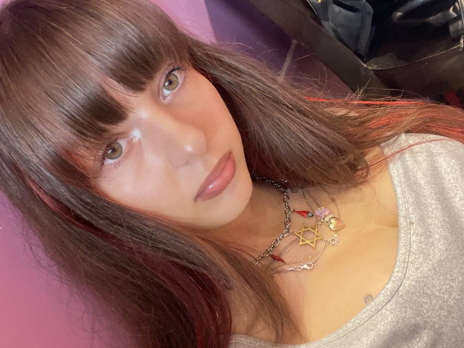 A woman with bangs and light brown hair with streaks of red smiles at the camera. Her hazel eyes are accentuated by makeup. She wears a gray shirt and multiple necklaces with various charms, including a Star of David and a heart. The background features a pink wall and a glimpse of black leather and possibly another person.