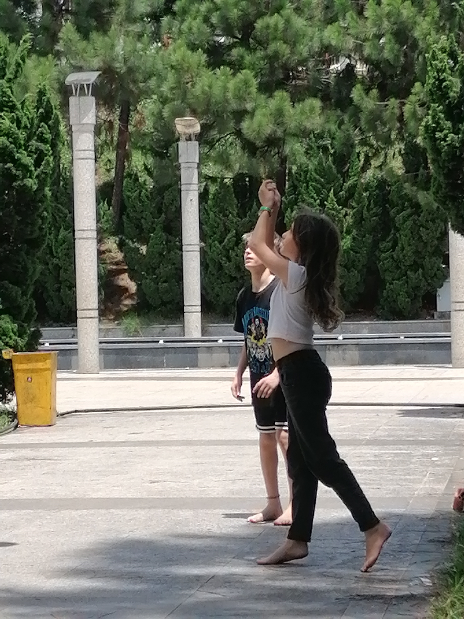 In Da Lat, Vietnam, two young people are seen outdoors. A girl in the foreground, wearing a white crop top and black pants, is barefoot and has her arms raised as if tossing something into the air. A boy, partially visible behind her and to the left, is also barefoot and appears to be looking up as well. He is wearing a black t-shirt with a graphic print. The setting is a paved area with trees and pillars in the background. A yellow trash bin is visible to the far left. The lighting suggests it is daytime, possibly late morning or early afternoon.