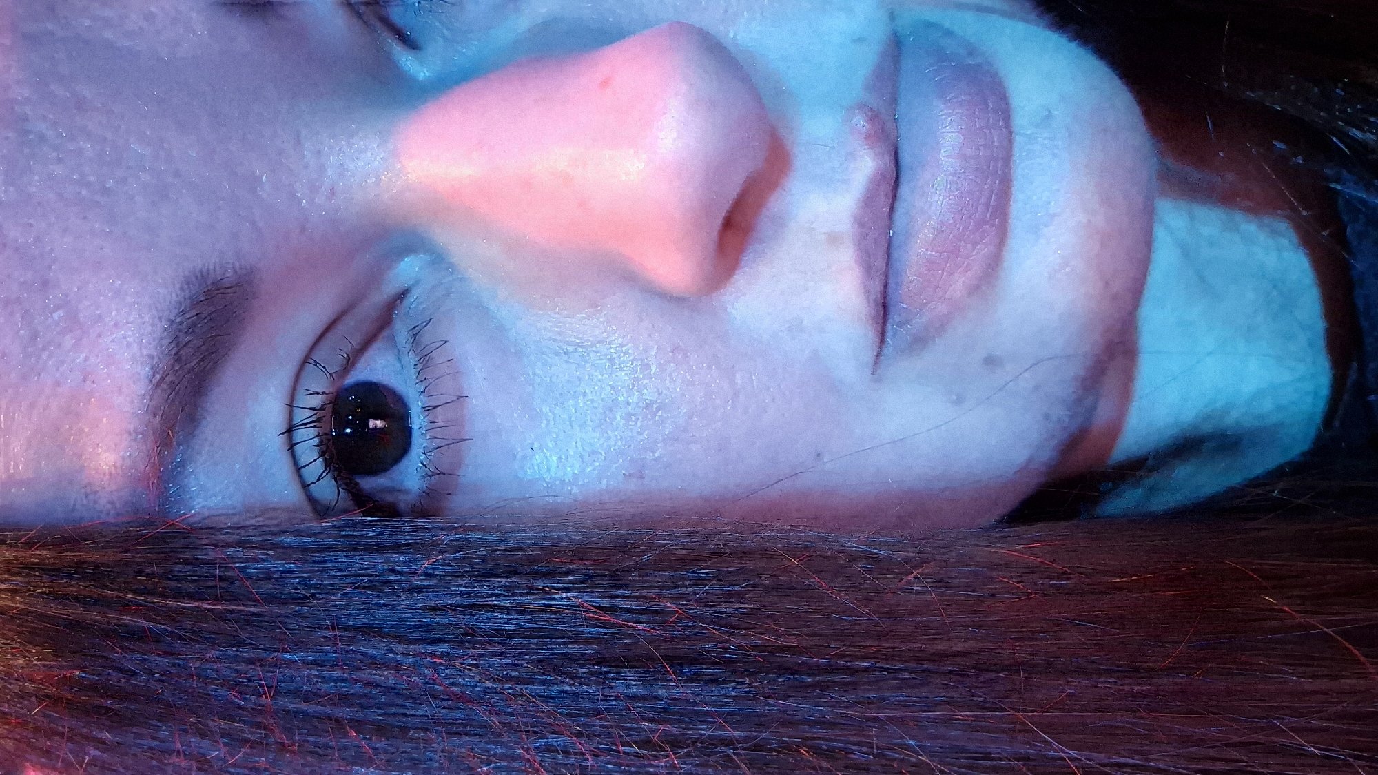 The image is a close-up, low-angle portrait of a person's face, focusing on the right eye, nose, and mouth. The subject's dark hair is visible on the left side of the frame. The lighting is dramatic, with strong blue and pink hues illuminating the skin, creating a somewhat ethereal or artificial atmosphere. The skin appears to have some texture and a slight sheen. The subject is looking directly at the camera. There are no discernible environmental details that would indicate a specific location in Kemerovo, Russia. The image appears to be taken indoors, possibly with colored lighting.