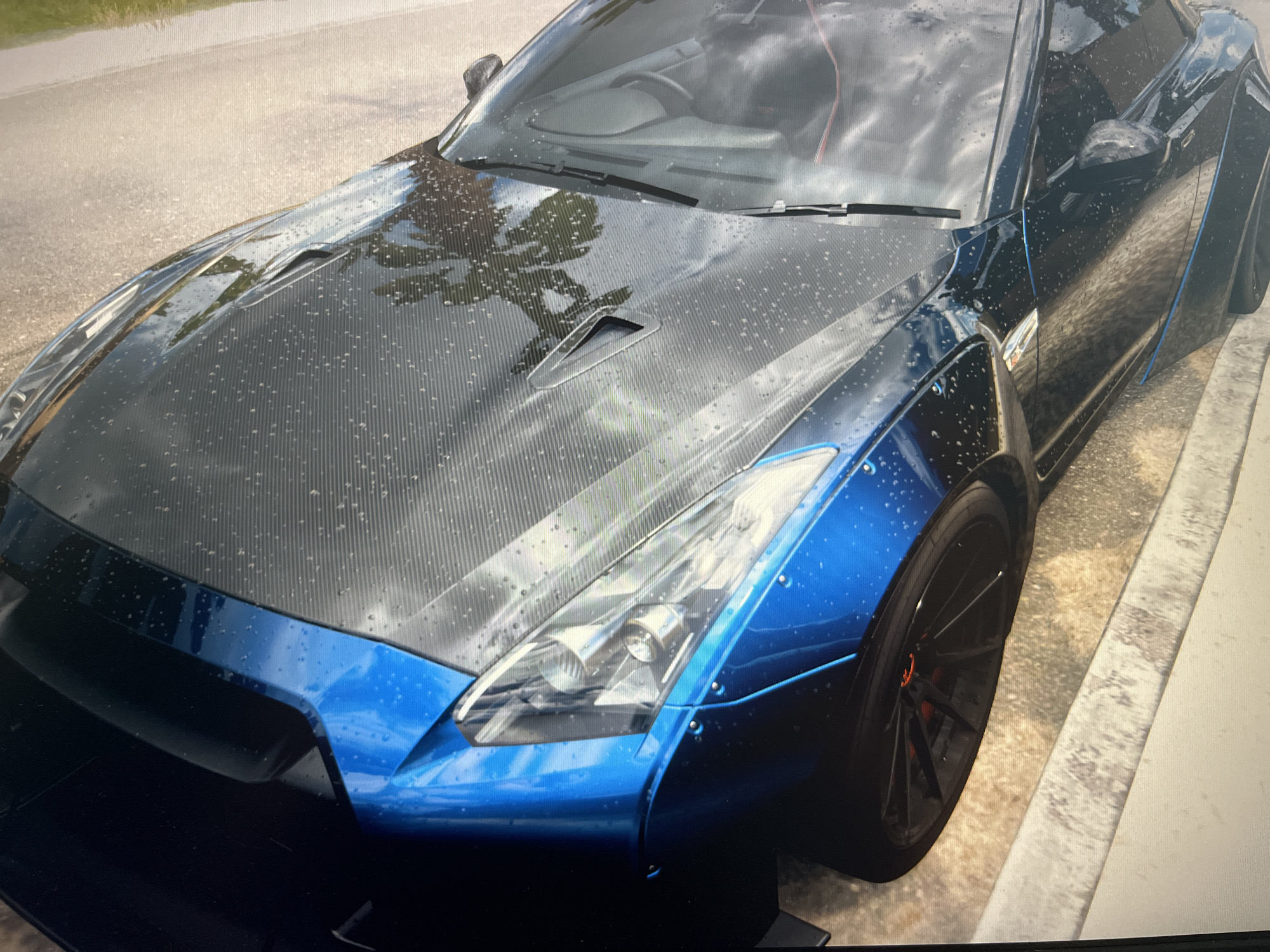 The image shows the front-left quarter of a dark blue sports car, possibly a Nissan GT-R, with a carbon fiber hood. The car is wet, with numerous small water droplets on its surface. The car is parked on the side of a road, with a concrete curb visible on the right side of the frame. The ground appears to be asphalt, with some gravel and dirt. The background is out of focus, but appears to be a natural environment with greenery and a body of water, possibly a lake or the sea. There are no people or any other discernible objects in the scene. The image is taken from a low angle, looking down at the car. The lighting suggests it is daytime, possibly overcast given the reflections on the car. There is no information to indicate this scene is in Varna, Bulgaria.