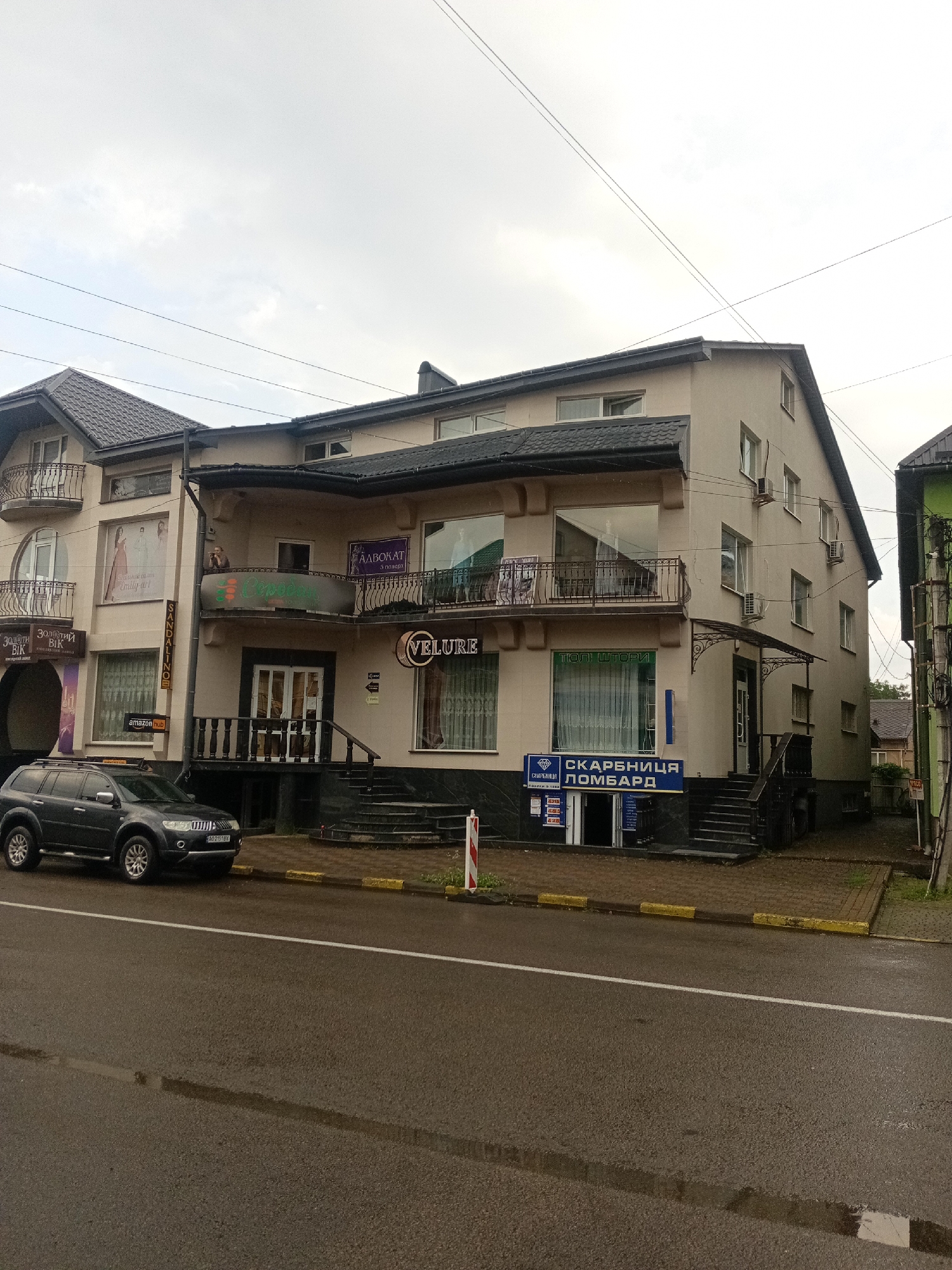 The image shows a storefront and a dark-colored SUV parked on the side of a wet asphalt road. The building is a multi-story structure with a beige facade. On the left side of the building, there are advertisements and balconies. A sign above a window reads "VELURE". Below this sign, there is a blue sign that says "СКАРБНИЦЯ ЛОМБАРД" (meaning pawn shop). This pawn shop appears to have several price listings displayed in its window. The street is wet, indicating recent rain, and there are yellow and grey curbs. The sky is overcast. The building is located in the city of Tiachiv, Ukraine.