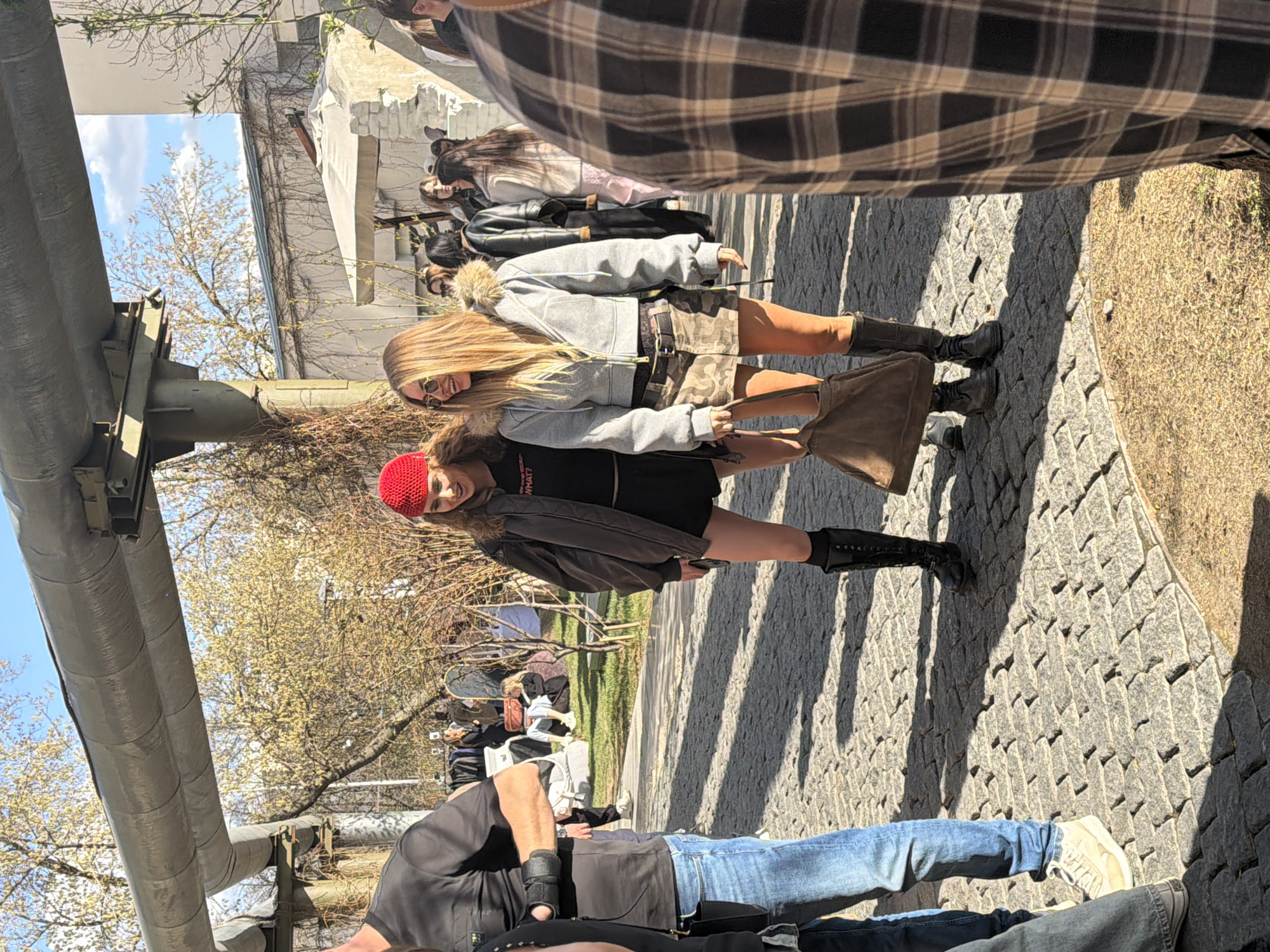 The image captures a street scene with several people walking. A pair of young women, one wearing a red beanie and the other with blonde hair, stand out in the foreground. They are wearing jackets and boots. One carries a brown bag. Other people can be seen further back.
The setting appears to be an urban environment, with cobbled pavement and trees lining the street. There are metal pipes running along the side of the road.
The city of Moscow, Russia, is not discernible from the image alone. There are no specific landmarks or characteristics in the photo to confirm the location. Therefore, it is impossible to determine with certainty whether it is in Moscow.